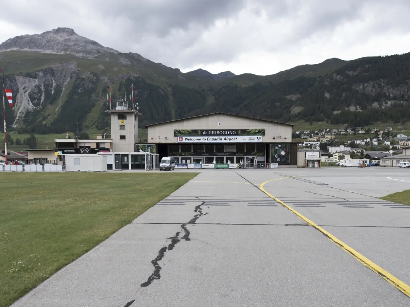 L'entrata dell'aeroporto di Engadin, con un edificio principale e una torre di controllo, circondato da montagne e vegetazione. Una pista di atterraggio visibile in primo piano.