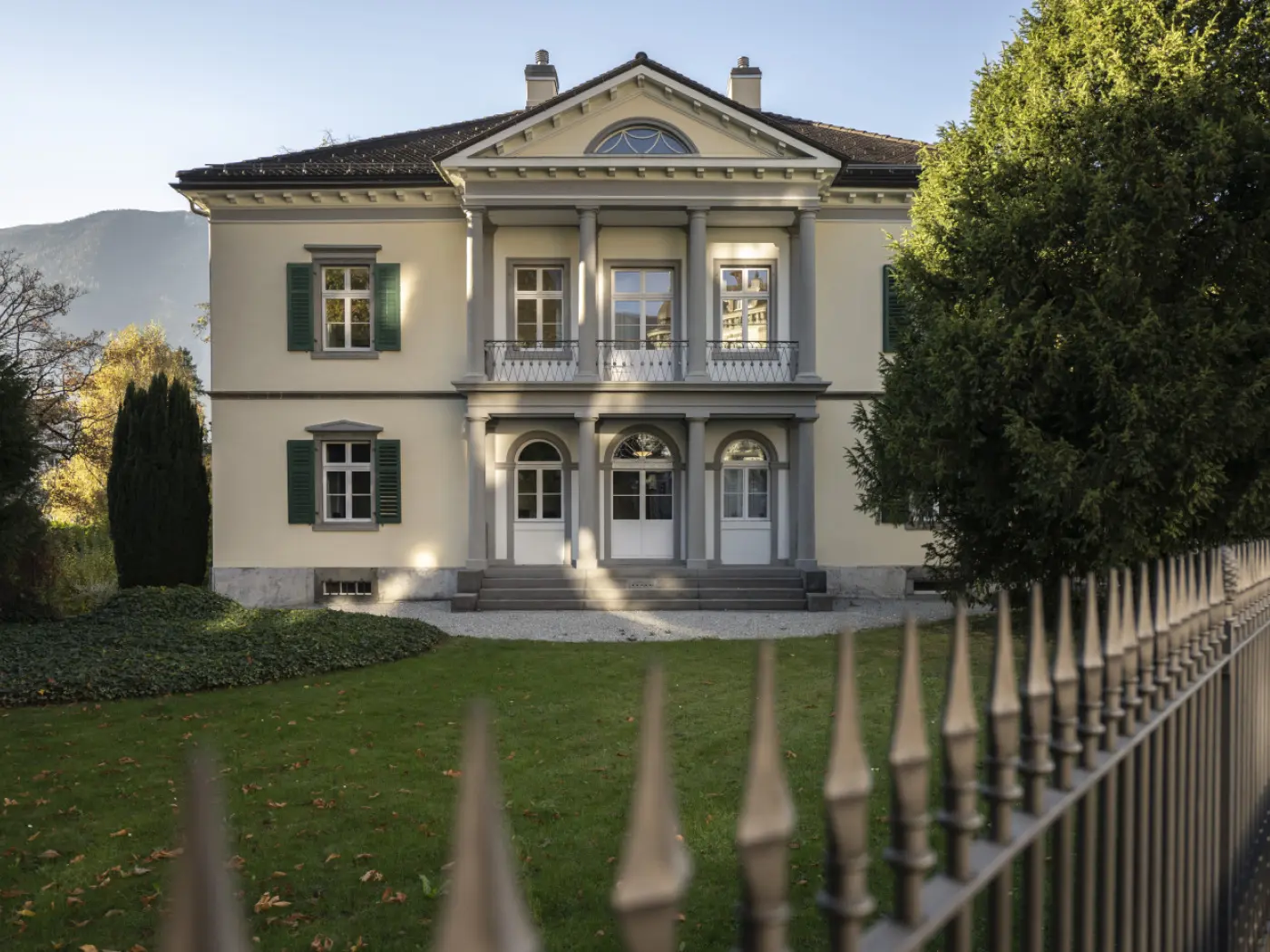 Villa elegante con facciata chiara e finestre verdi. Il giardino è curato, con alberi e una recinzione in ferro battuto. La montagna si intravede sullo sfondo.