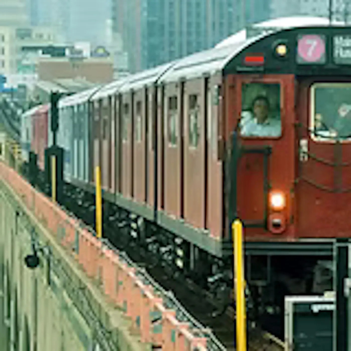 Treno della metropolitana che percorre una sopraelevata, con un conducente visibile nel vagone. Sullo sfondo si intravedono edifici di una città.