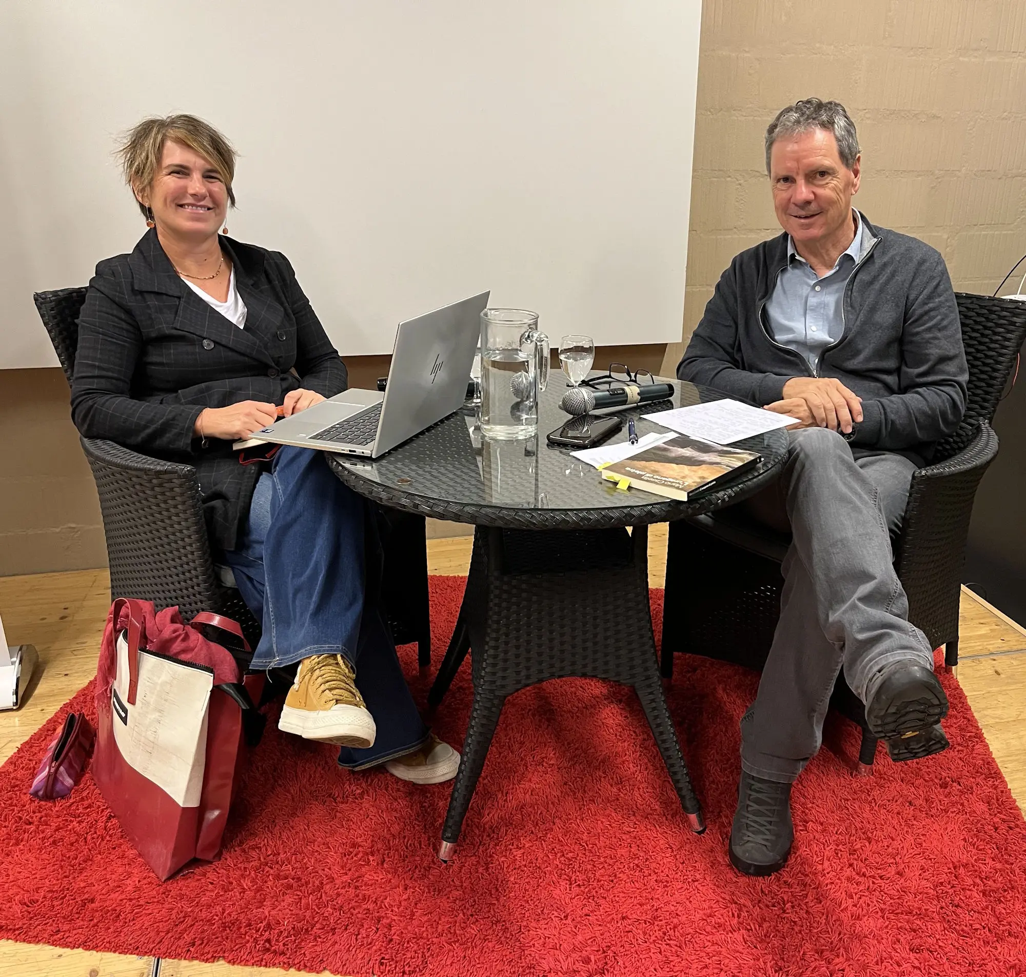 Due persone sedute attorno a un tavolo, con computer portatile e documenti. Una donna con capelli corti e giacca nera, e un uomo con maglione grigio, entrambi sorridenti. Sul tavolo ci sono una bottiglia d'acqua e un registratore. Il pavimento è coperto da un tappeto rosso.
