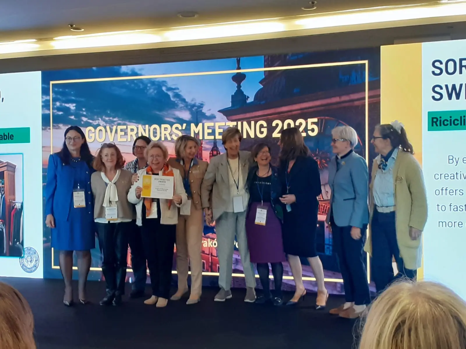 Gruppo di donne sorridenti sul palco durante un incontro dei governatori, con un grande schermo sullo sfondo che mostra "Governors' Meeting 2025". Una di loro tiene un certificato.