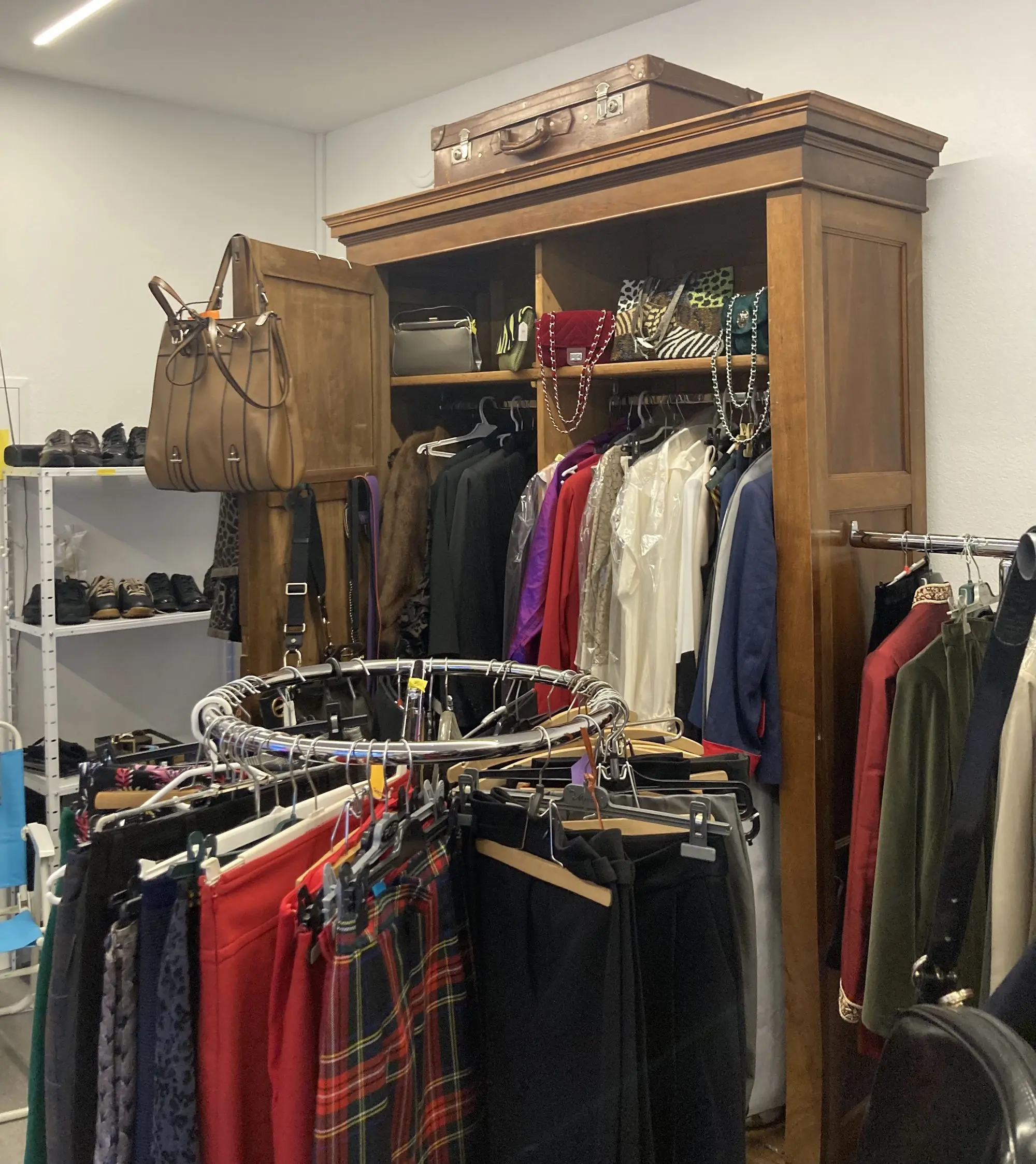 Armadio di legno con vestiti appesi, accanto a una struttura circolare con gonne e pantaloni. Un handbag è appeso a sinistra e una valigia è posizionata in cima all'armadio. La scena è in un ambiente ben illuminato.