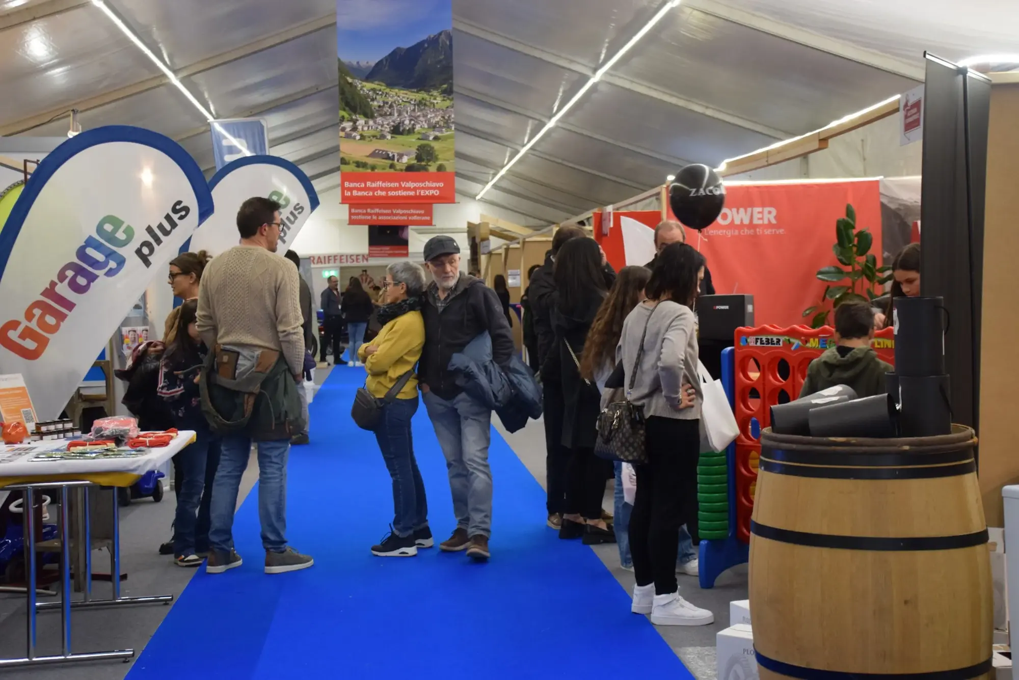 Folla di visitatori in una fiera, con stand decorati e un tappeto blu. Alcune persone discutono, mentre altri osservano prodotti esposti. Sullo sfondo, poster e banner promozionali.