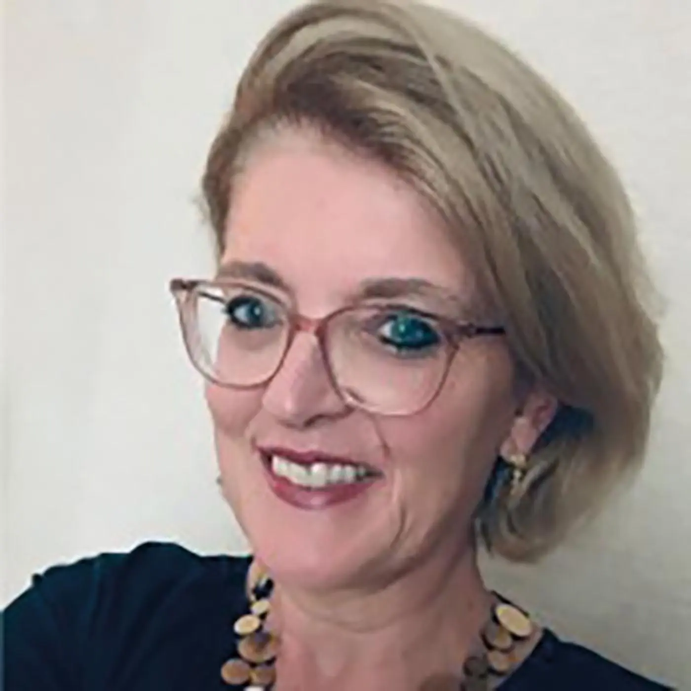 Donna con capelli biondi e occhiali, sorridente e con collana di perline. Sullo sfondo si distingue una parete chiara.