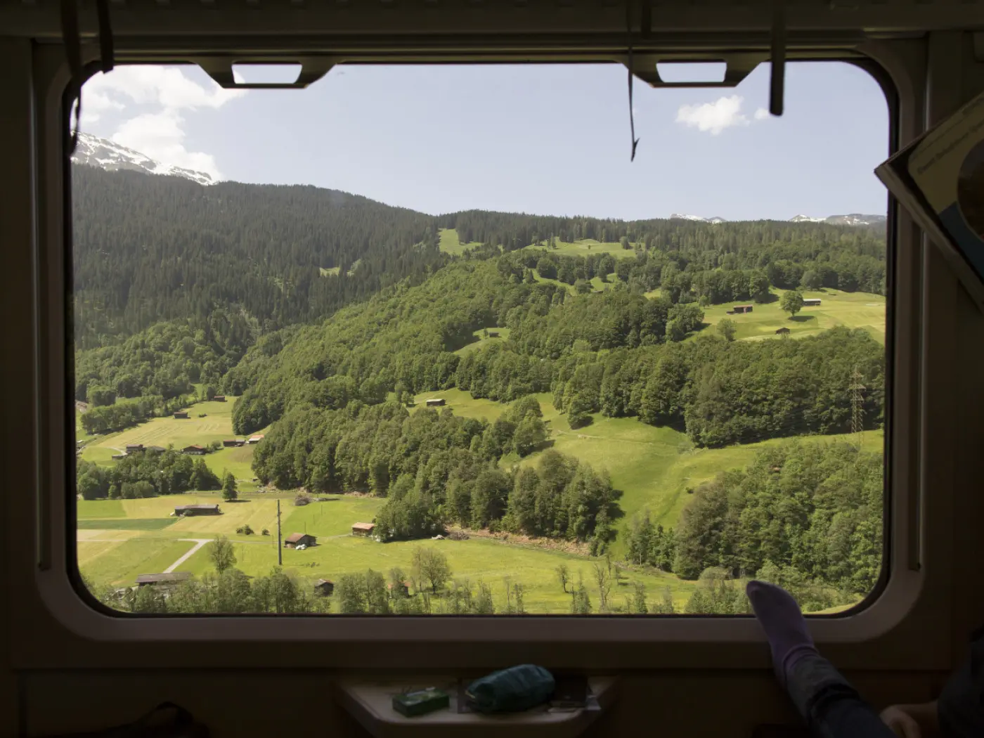 Vista panoramica da un treno con colline verdi ricoperte di alberi e prati, sotto un cielo parzialmente nuvoloso.