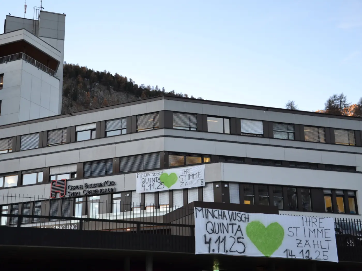 Edificio con striscioni affissi all'esterno. I cartelli mostrano scritte in nero e verde, con date e frasi come "MINCHA WUSCH QUINTA 4.11.25" e "IHRE STIMME ZAHLT 14.10