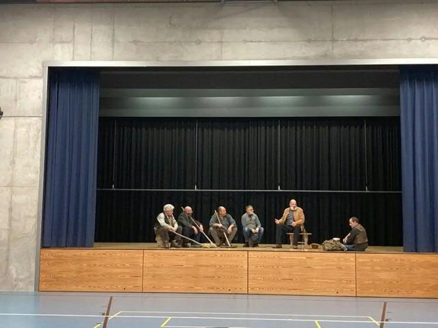 Un gruppo di uomini seduti su un palco di legno in un ambiente interno. Indossano abiti semplici e sono intenti a conversare. Sullo sfondo, tende nere coprono il muro.