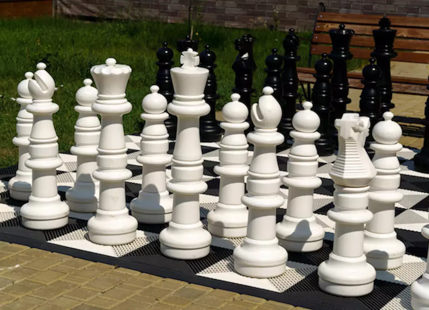 Scacchiera all'aperto con pezzi di scacchi bianchi e neri di grandi dimensioni. Sullo sfondo, una panchina di legno e un prato verde.