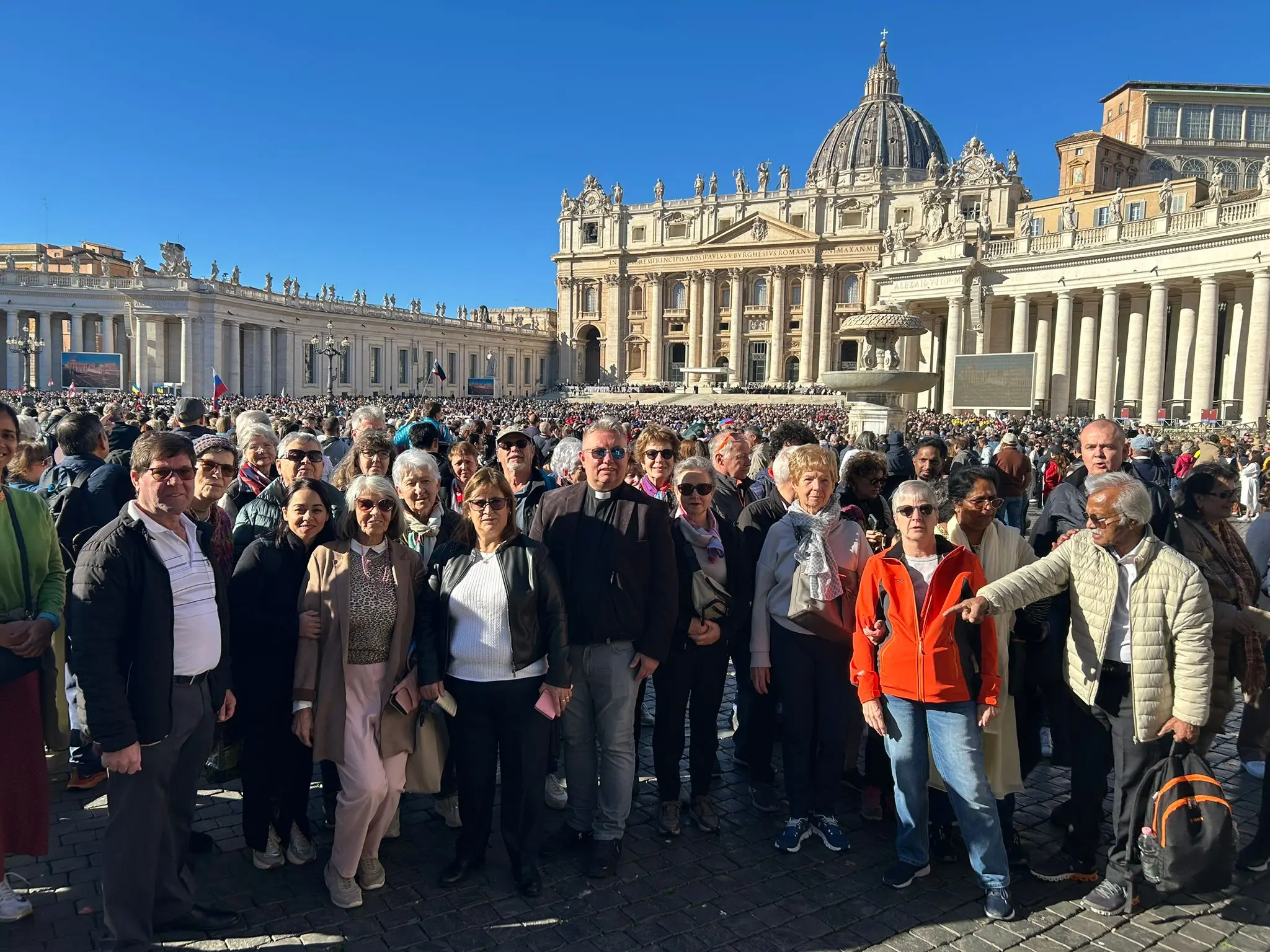 Un gruppo di persone in piedi nella piazza San Pietro, con la basilica e i colonnati sullo sfondo. È una giornata di sole, e numerosi visitatori si sono radunati per un evento.