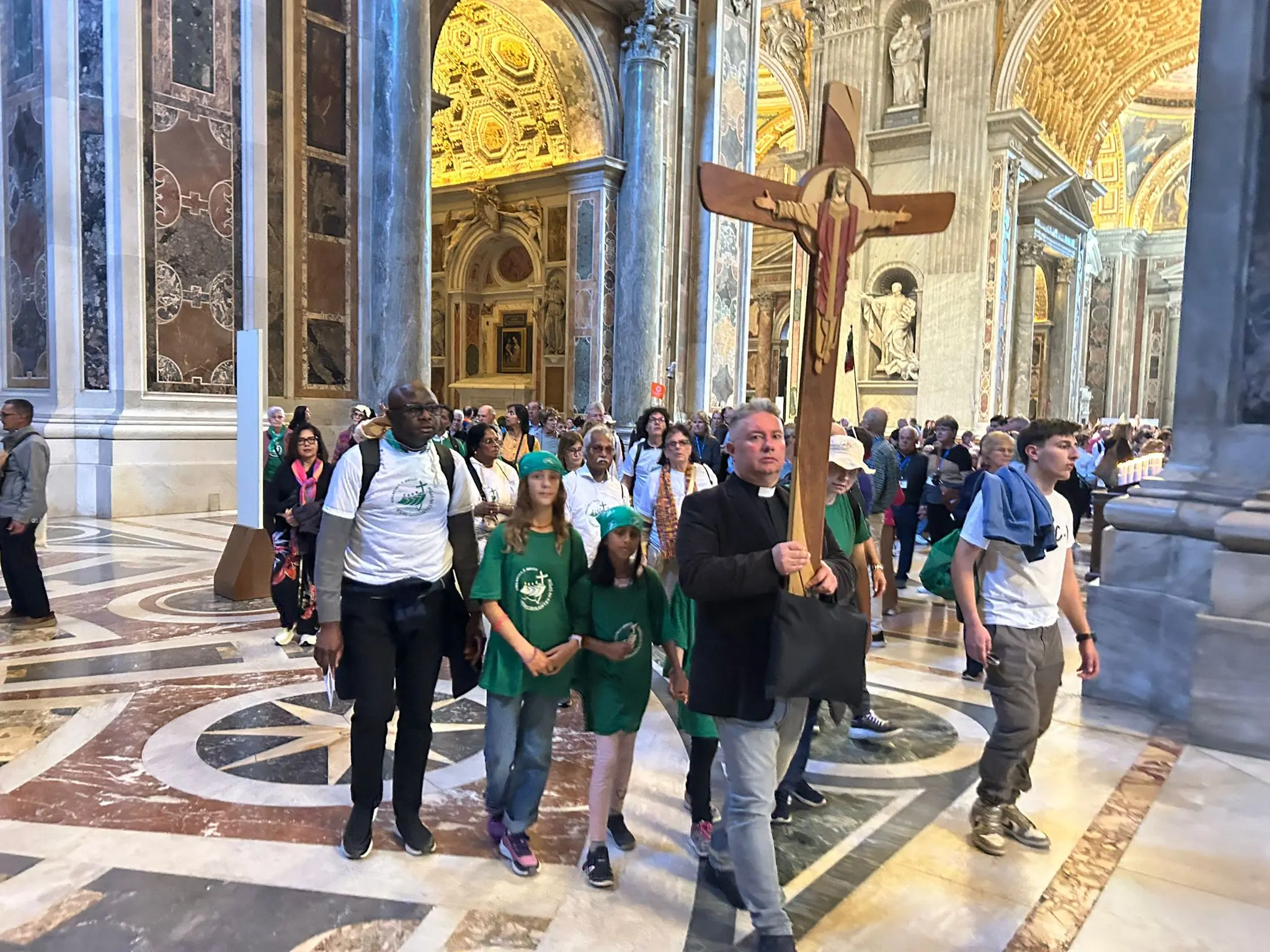 Gruppo di persone che cammina all'interno della Basilica di San Pietro, con un sacerdote che tiene una grande croce. L'illuminazione interna è calda e decorativa, con dettagli architettonici in evidenza.