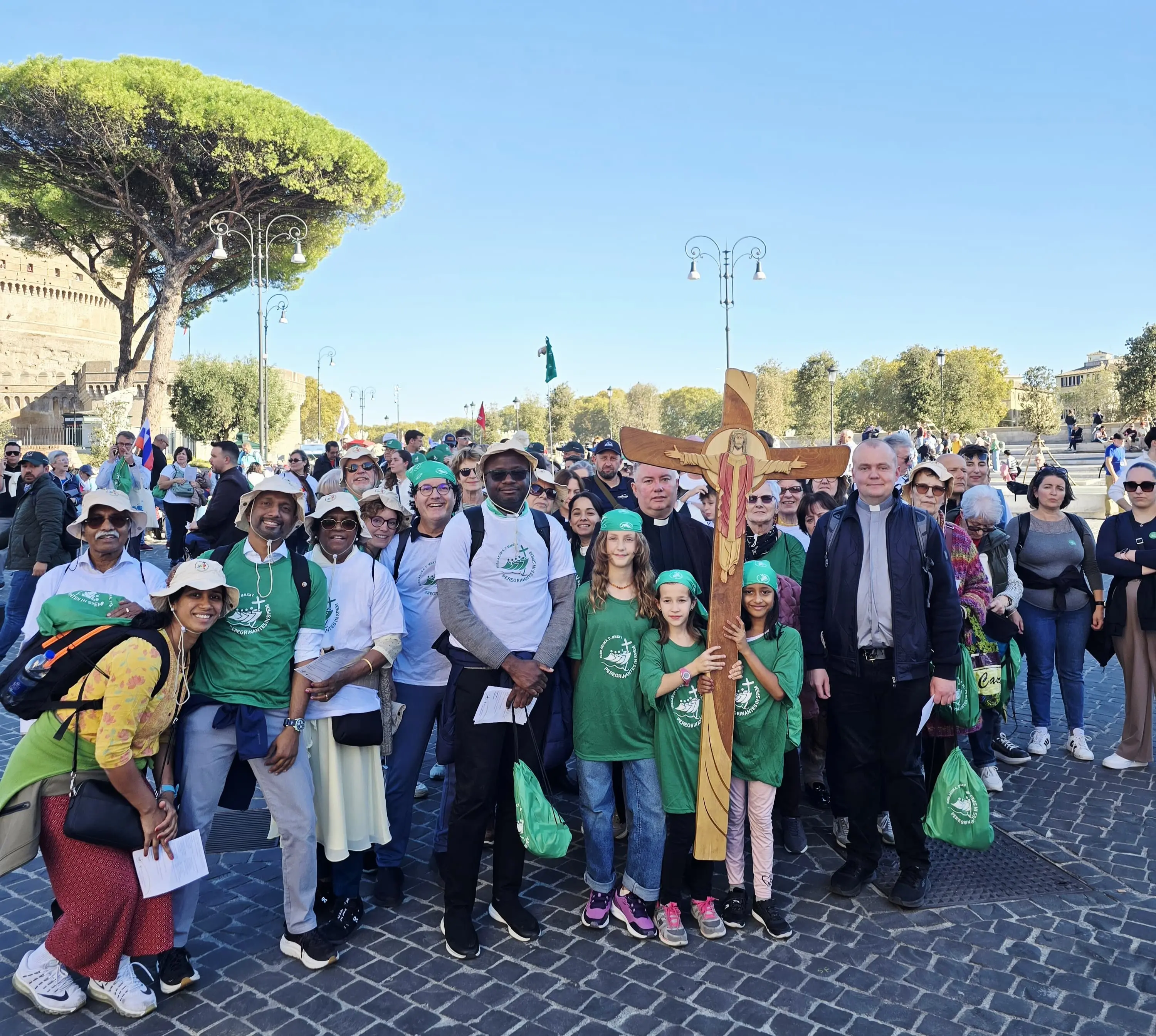 Un gruppo di persone sorridenti in magliette verdi si riunisce in una piazza, con alcuni che tengono una grande croce di legno. Sullo sfondo si vedono alberi e edifici storici.