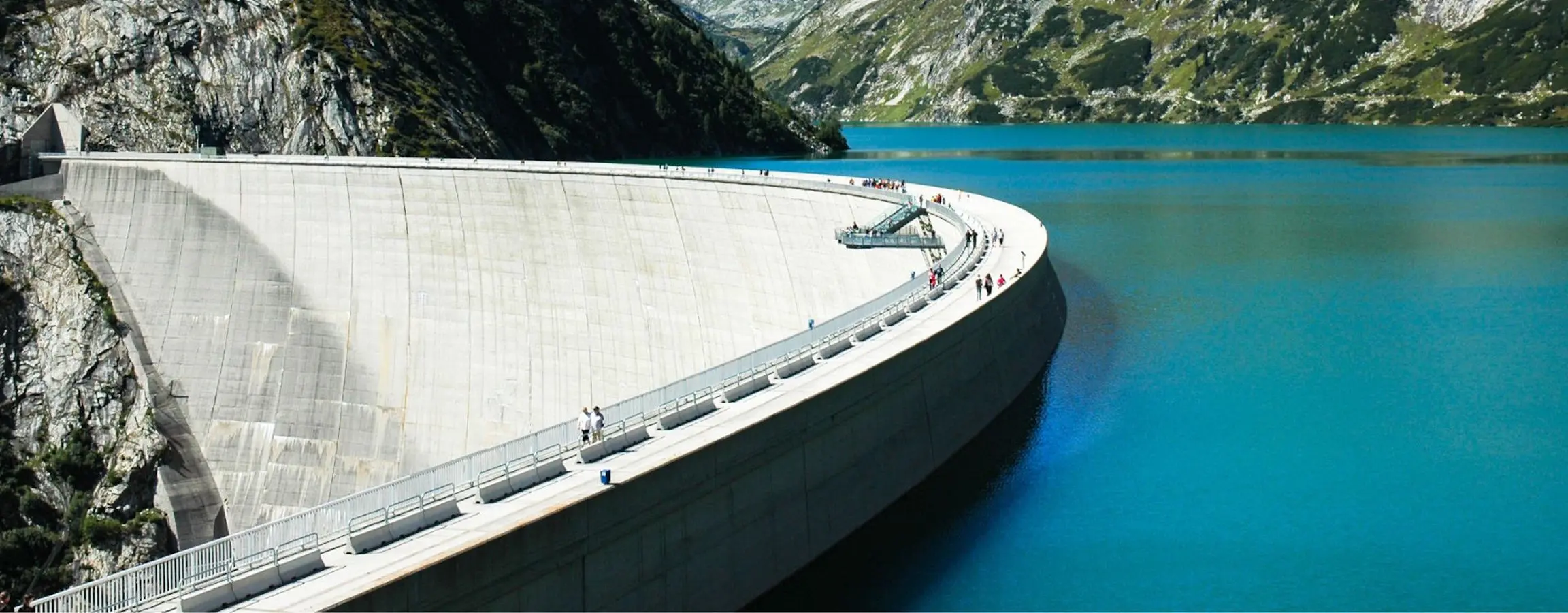 Diga circolare in cemento con una vista panoramica su un lago blu intenso e montagne verdi sullo sfondo. Persone passeggiano lungo il bordo della diga.