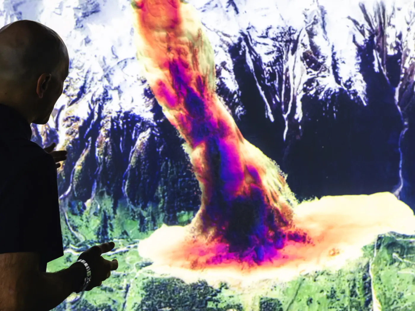 Un uomo osserva un'illustrazione digitale di un'eruzione vulcanica che emette un rivolo di lava e cenere, circondato da montagne e vegetazione.