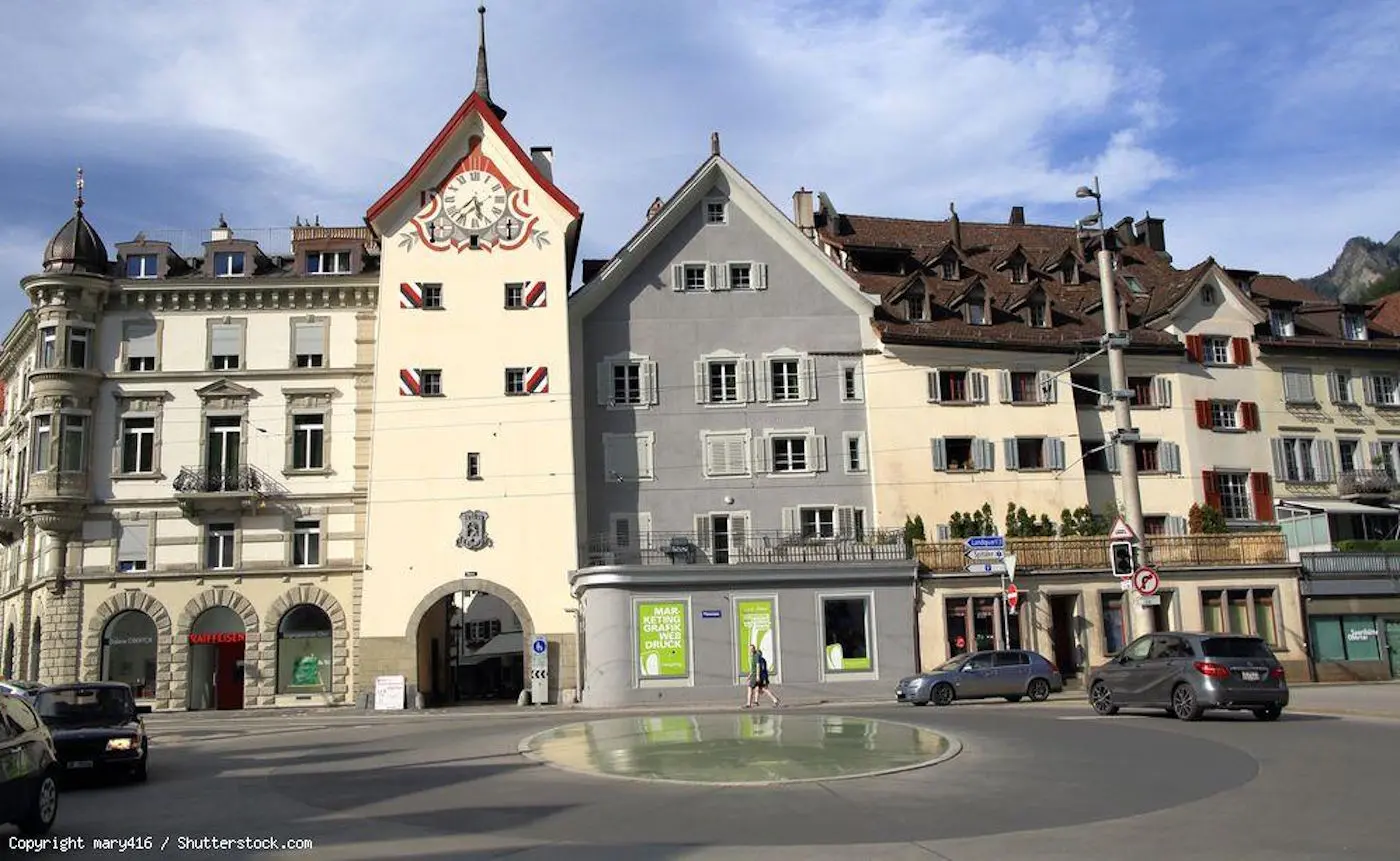 Una piazza con un edificio storico che presenta un orologio e decorazioni architettoniche. Al lato ci sono edifici moderni e un'area verde attorno a una fontana. Il cielo è parzialmente nuvoloso e si vedono altre strutture in lontan