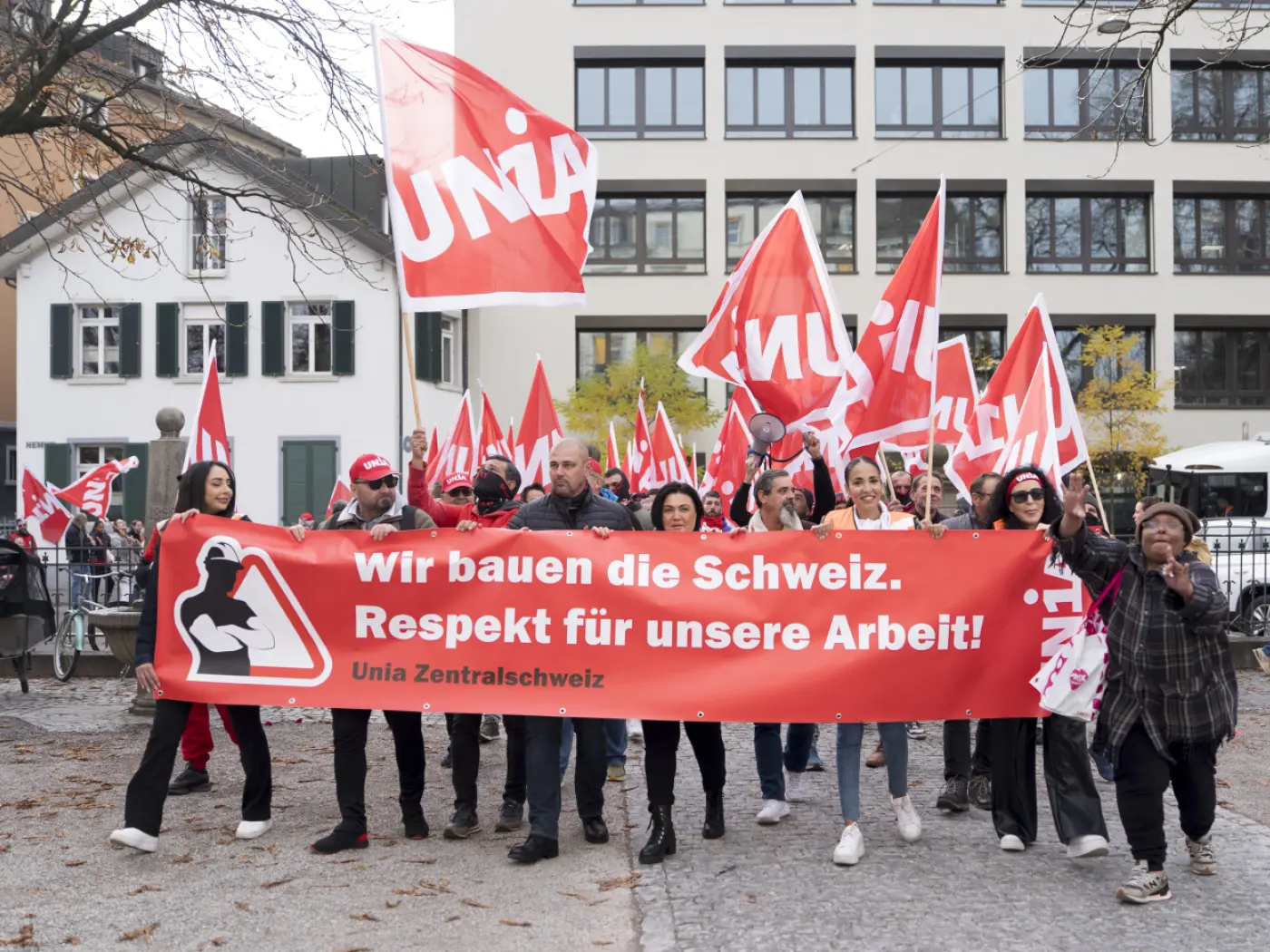 In una manifestazione, un gruppo di persone tiene bandiere rosse e uno striscione con la scritta in tedesco "Wir bauen die Schweiz. Respekt für unsere Arbeit!" mentre camminano in una zona urbana, circondati da edifici e alberi.