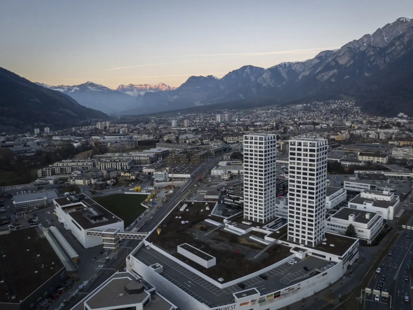 Vista aerea di una città con due grattacieli moderni e alti, circondati da edifici più bassi. Sullo sfondo si vedono montagne con cime innevate, mentre il cielo si tinge di un delicato colore al tramonto.