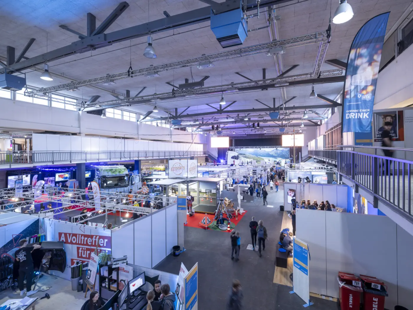 Fiera affollata con vari stand e esposizioni. Persone si muovono tra le aree, mentre lampade e schermi illuminano l'ambiente. Banner e pubblicità sono visibili lungo lo spazio.