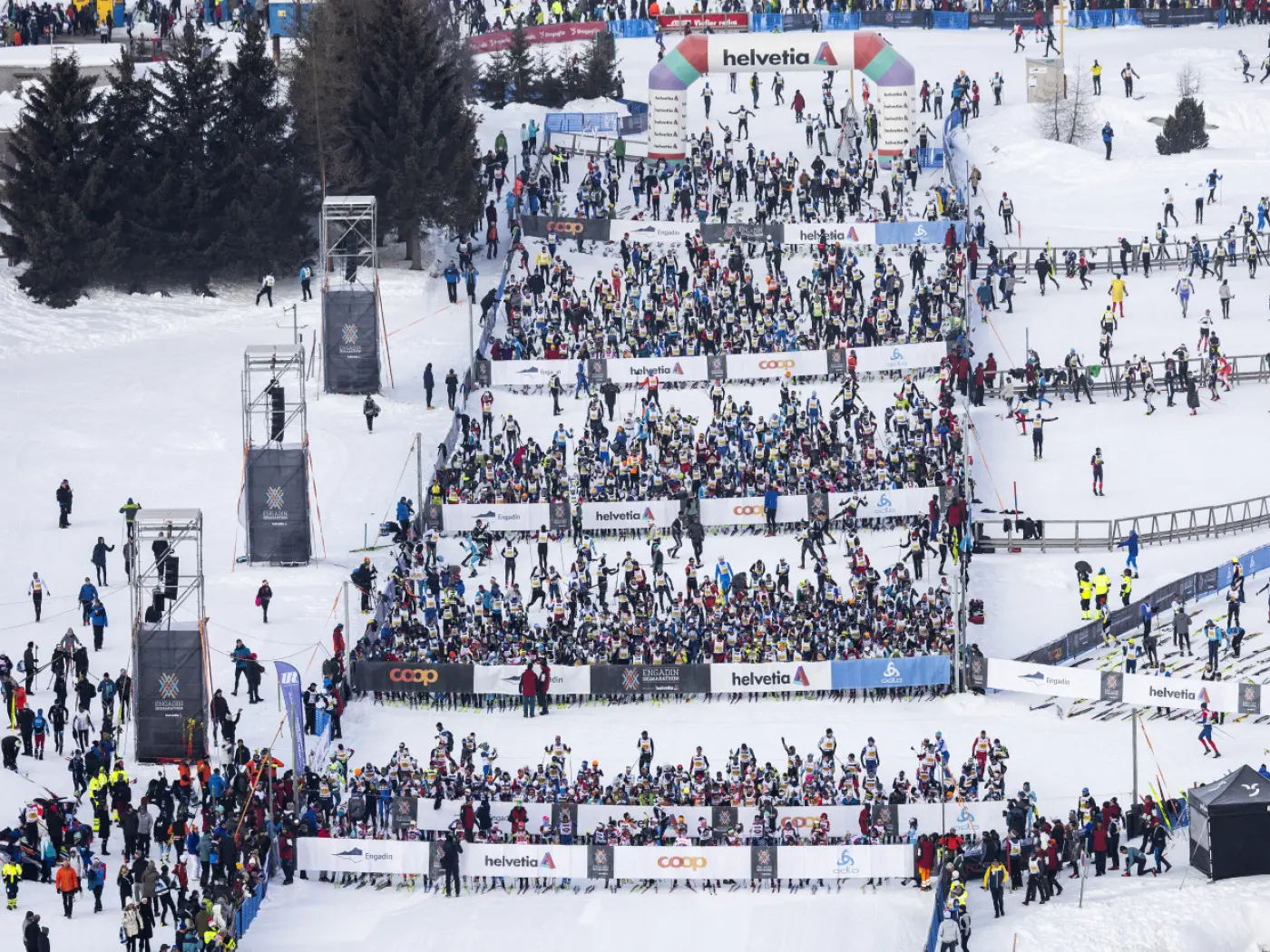 Tantissimi sciatore si preparano alla partenza in una località montana innevata, con bandiere e striscioni sulla neve. Ci sono diverse persone sugli spalti che assistono all'evento.