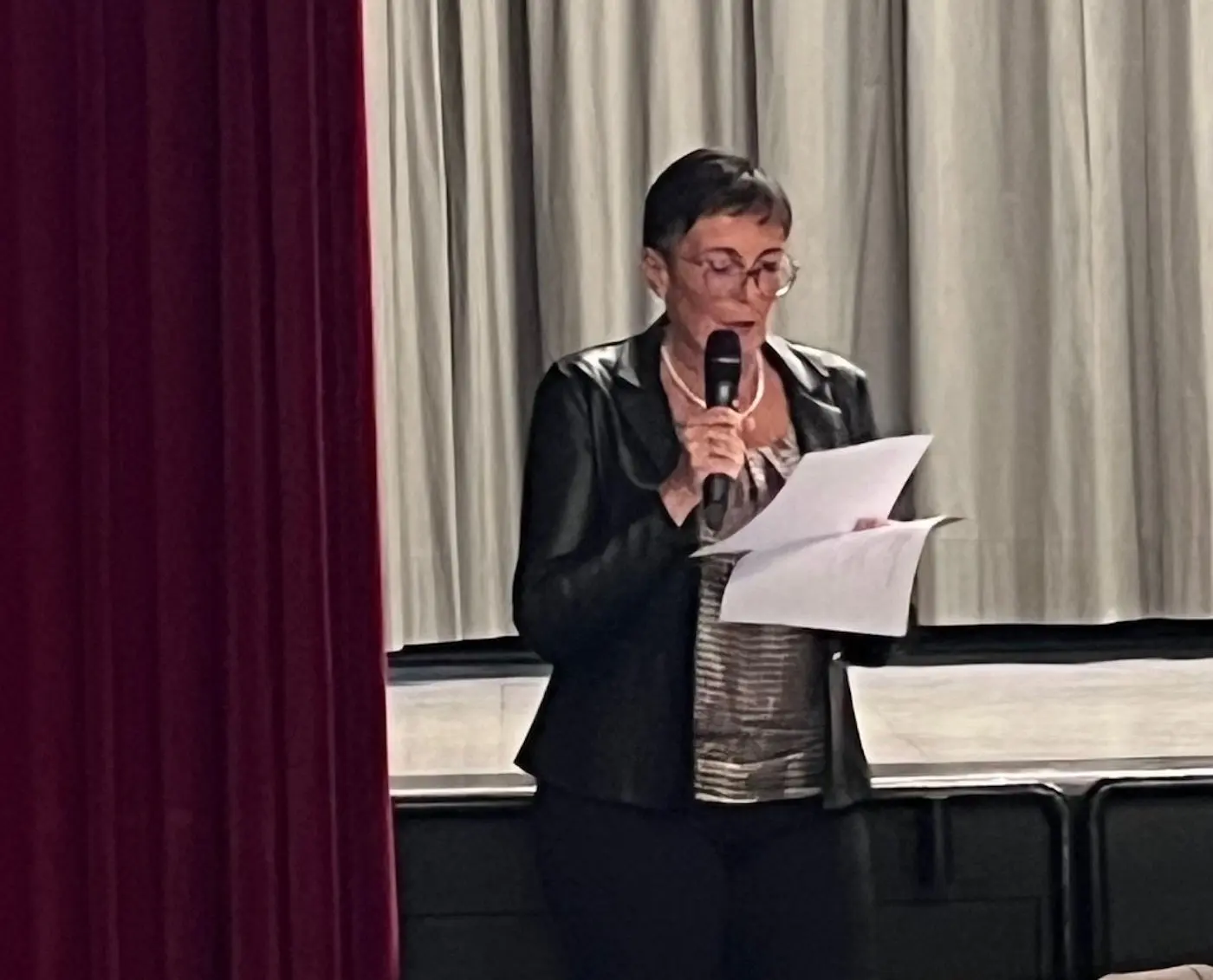 Una donna di mezza età con occhiali e un blazer nero sta leggendo un documento con un microfono in mano. Si trova su un palco con tende grigie sullo sfondo.