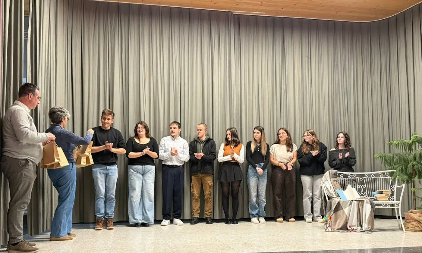 Un gruppo di giovani in piedi su un palco durante un evento, applaudendo. Alcuni stanno ricevendo premi o riconoscimenti mentre altri osservano. Sullo sfondo, una parete di tende e un mobile decorativo.