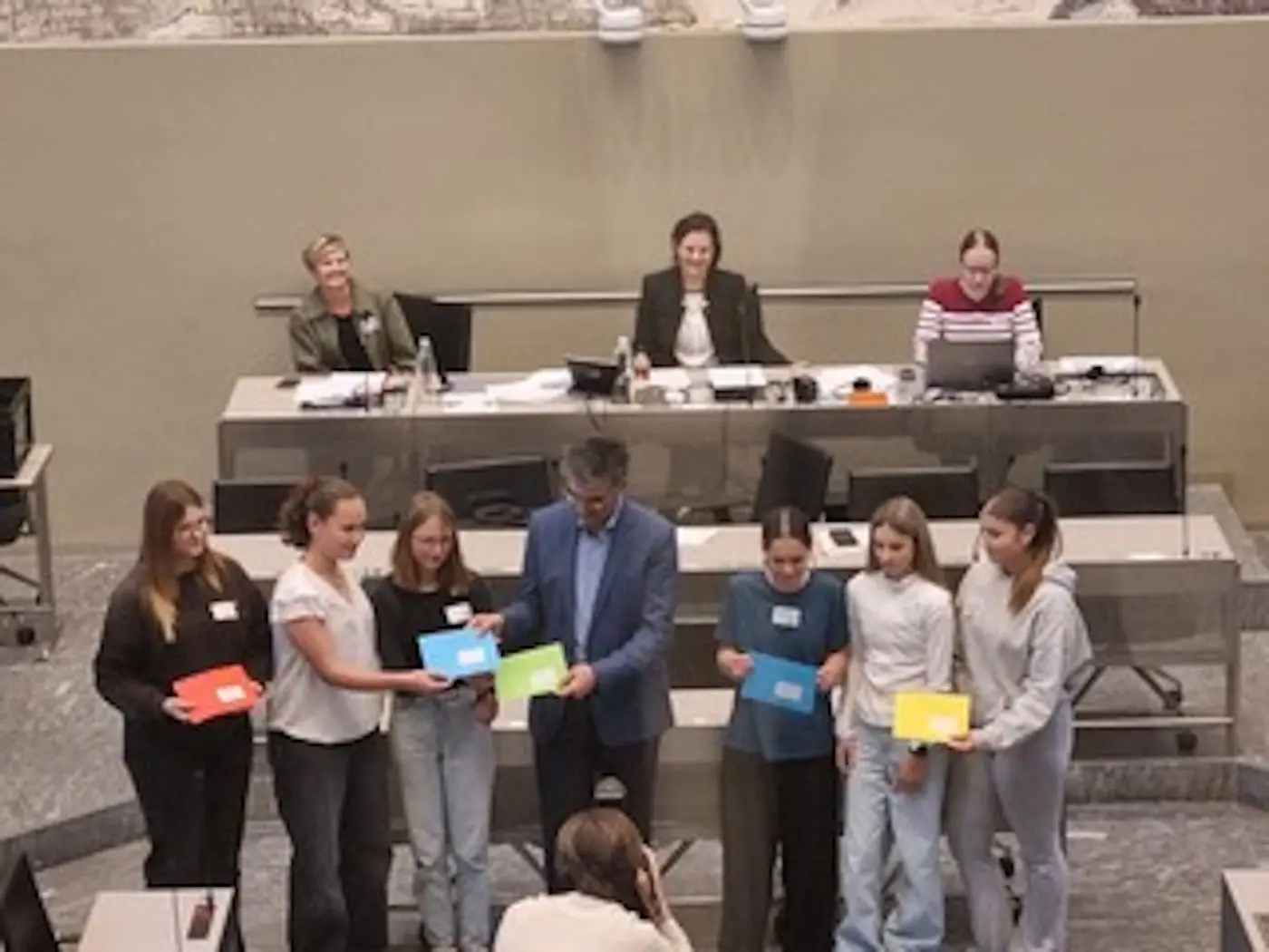Sei ragazzi ricevono riconoscimenti durante una cerimonia in un'aula di tribunale. Un uomo al centro consegna delle buste colorate, mentre gli altri sorridono. Sullo sfondo, alcune donne assistono all'evento, sedute al tavolo.