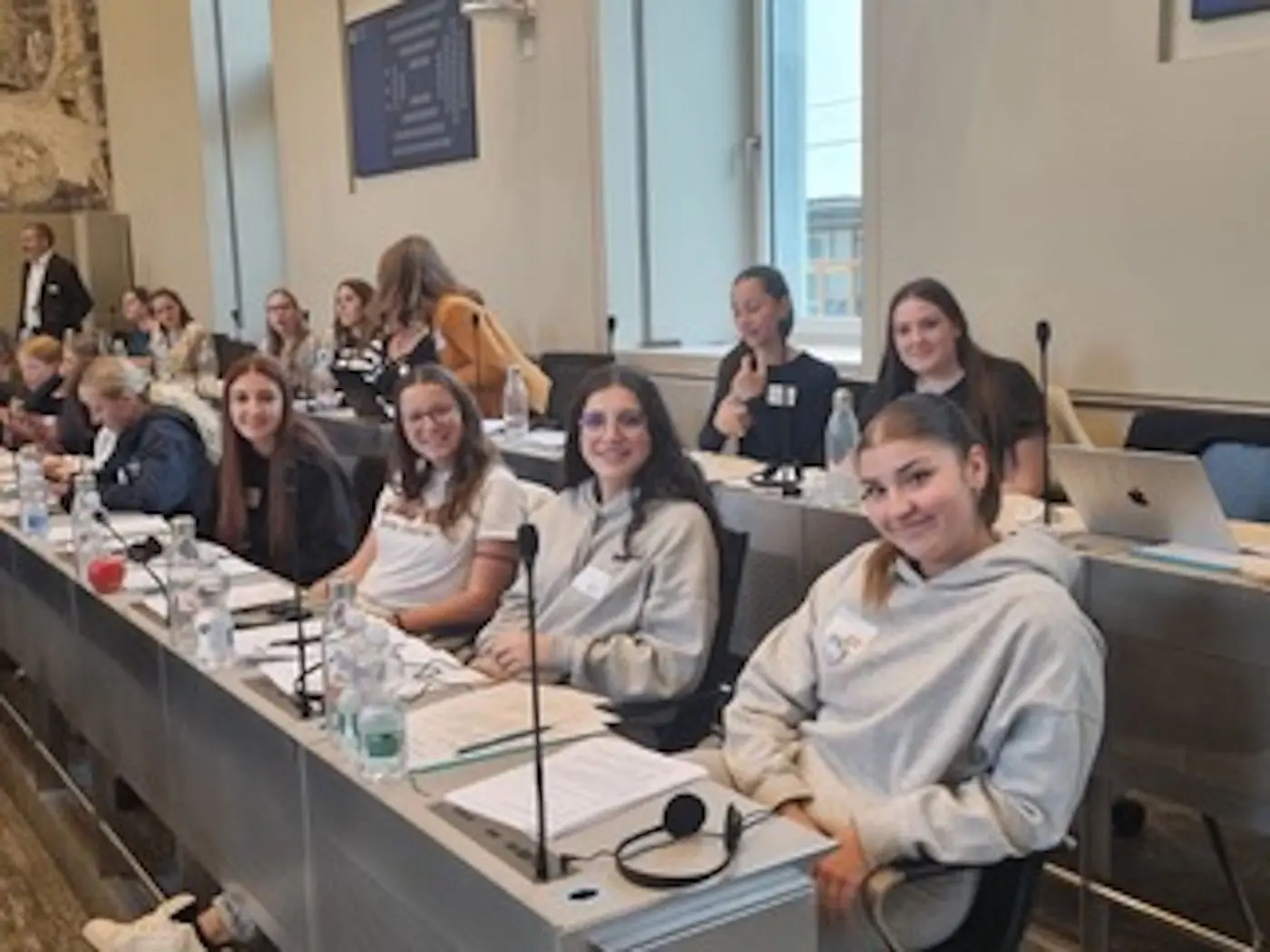 Un gruppo di sei ragazze sedute a un tavolo in una sala conferenze. Alcune di loro sorridono mentre altre sono concentrate, con laptop e bottiglie d’acqua sul tavolo. Il muro dietro di loro presenta decorazioni e finestre che lasciano entrare