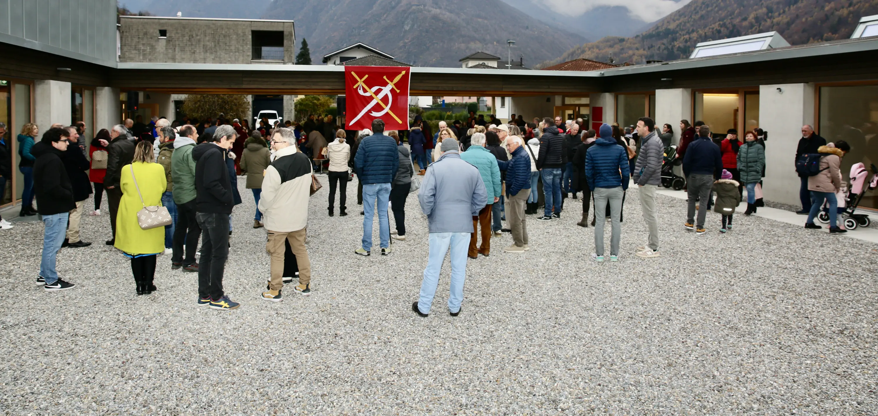 Un gruppo di persone radunate in un'area all'aperto, con un'ambientazione montana sullo sfondo. Alcuni indossano giacche invernali e ci sono bandiere rosse visibili.
