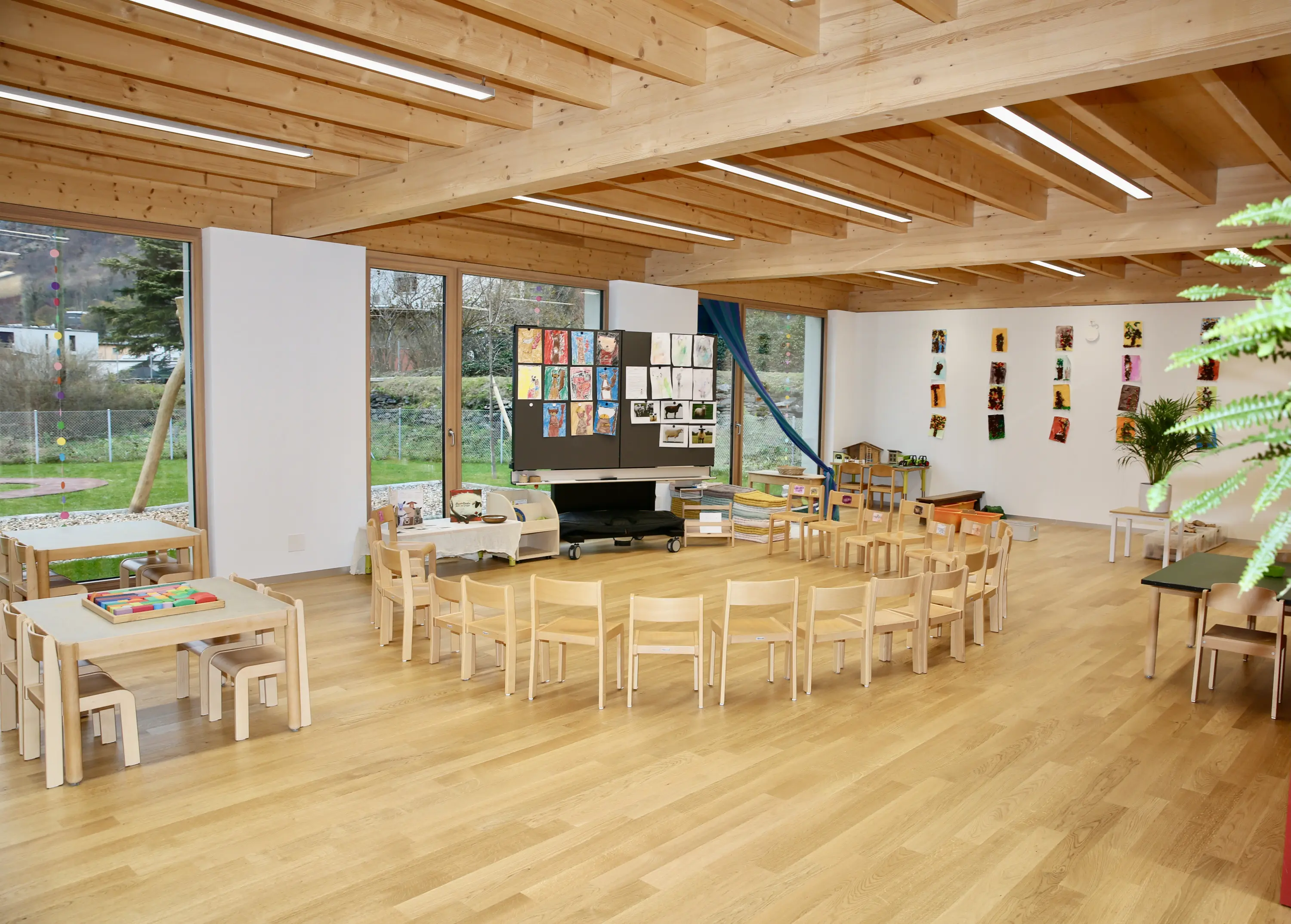 Aula scolastica moderna con pareti bianche e travi di legno a vista. Tavoli e sedie di legno chiaro disposti in cerchio, con un'area dedicata all'arte e opere esposte sulle pareti. Una grande finestra offre luce naturale