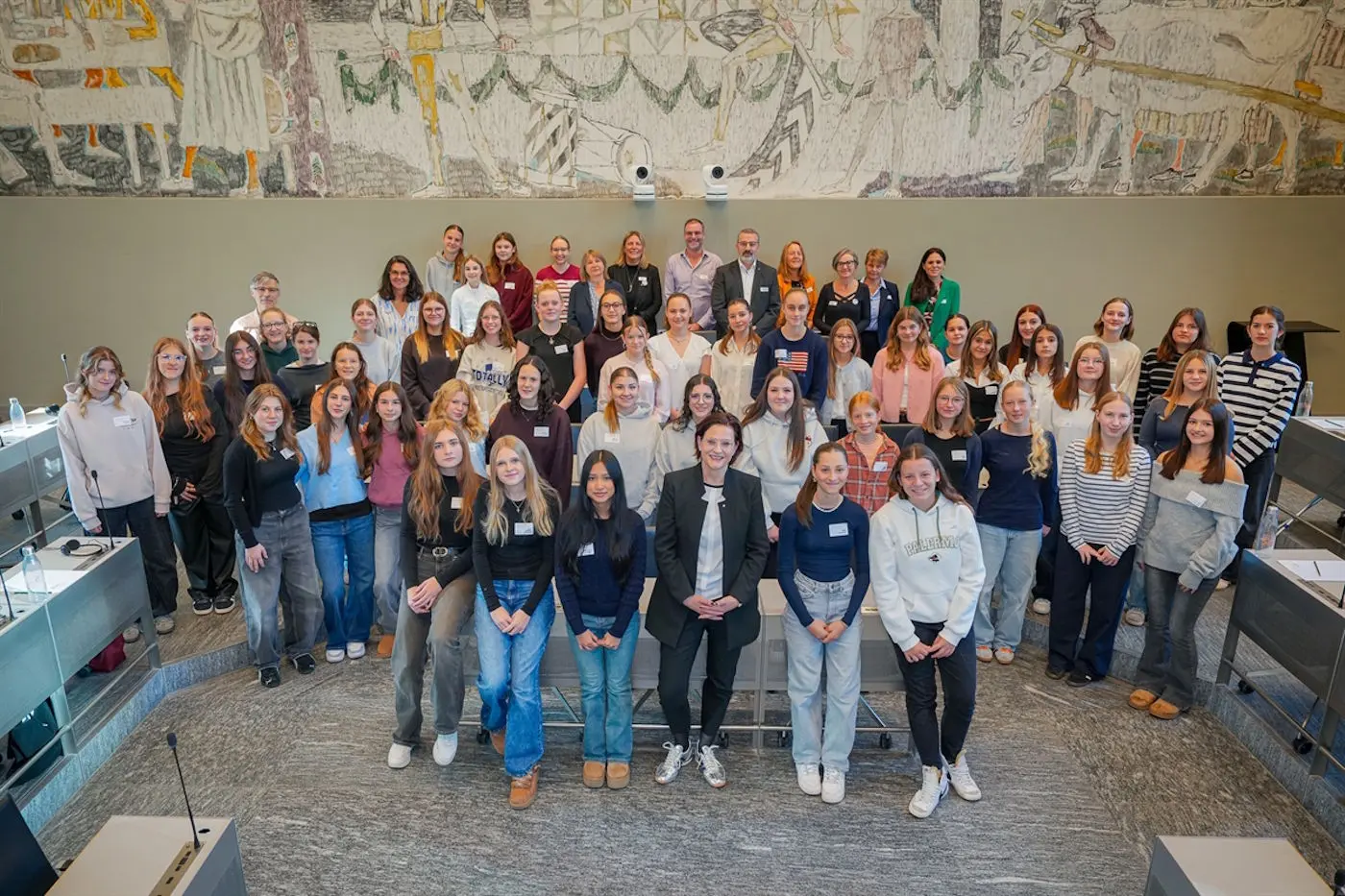 Gruppo di circa 60 persone, principalmente donne, riunite in un ambiente ufficiale con scrivanie e un grande arazzo sullo sfondo. Tutti sorridono e sono vestiti in modo casual.