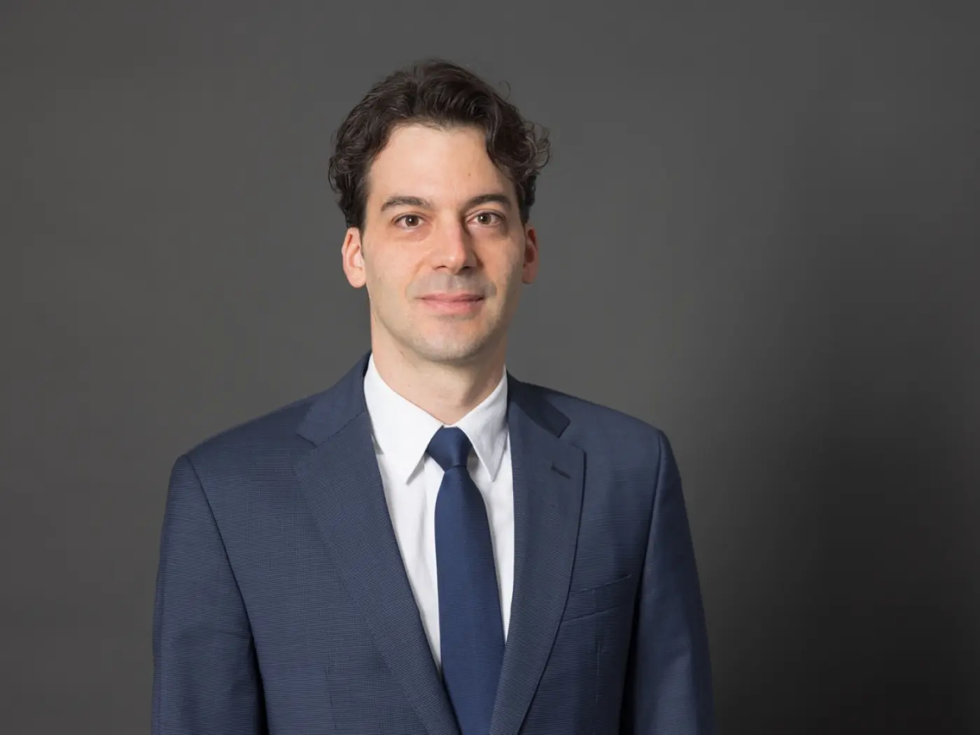 Uomo con capelli scuri e ricci, vestito in un completo blu, cravatta blu scuro, posa di fronte a uno sfondo grigio.