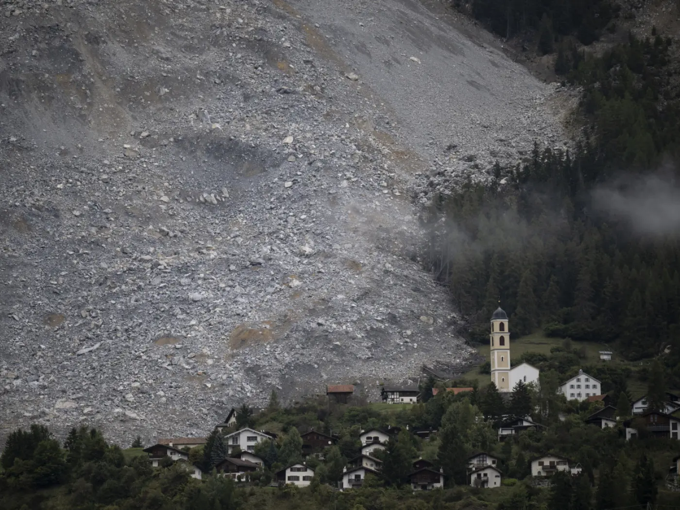 Un'ampia frana si è staccata da una montagna, circondando un piccolo paese con una chiesa e diverse case. Sullo sfondo si vedono alberi e nuvole.