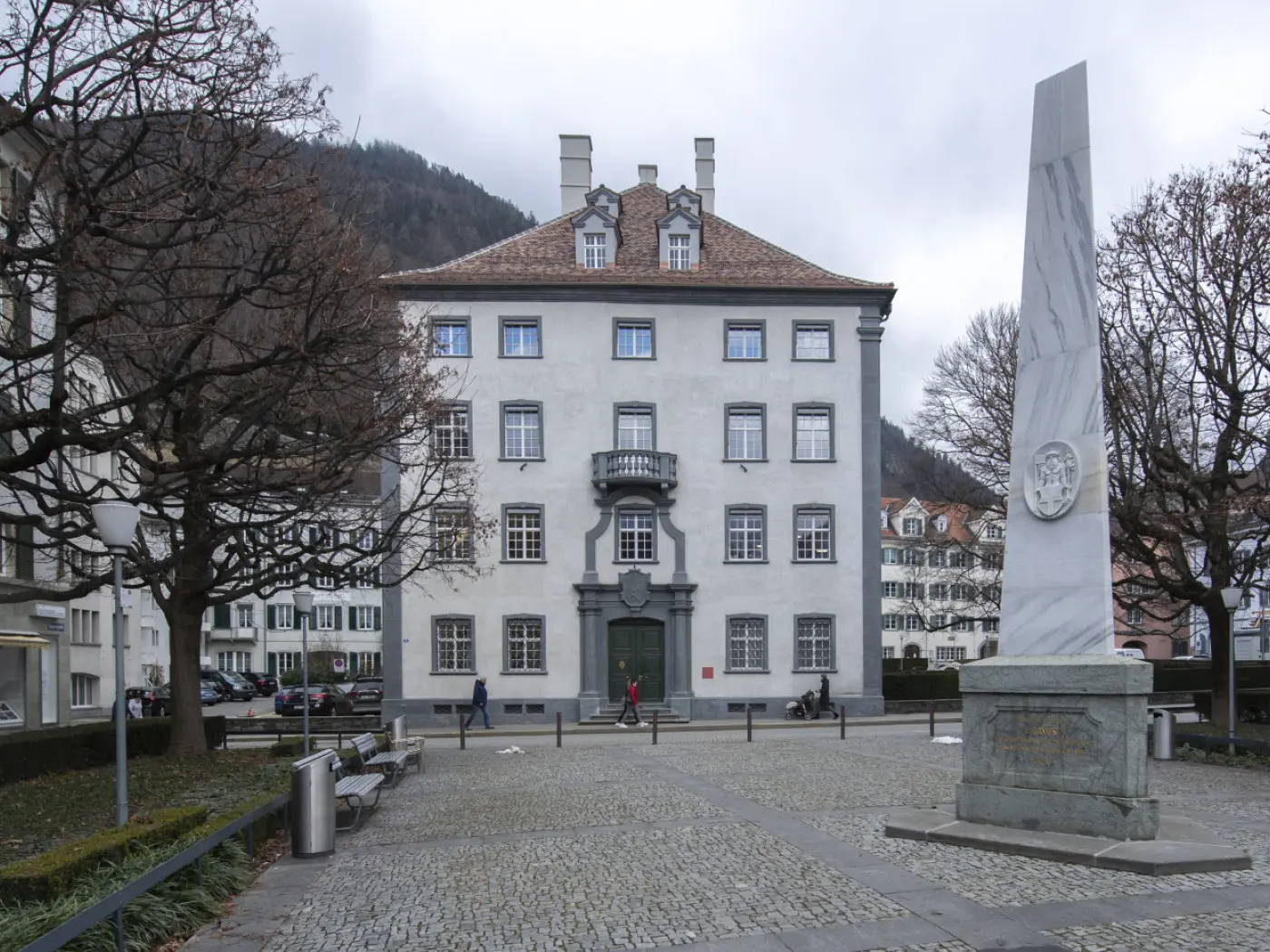 Un edificio storico con facciata grigia e tetto rosso in una piazza alberata. Un monumento di pietra si trova di fronte all'edificio. Il cielo è nuvoloso e ci sono panchine e persone nella scena.