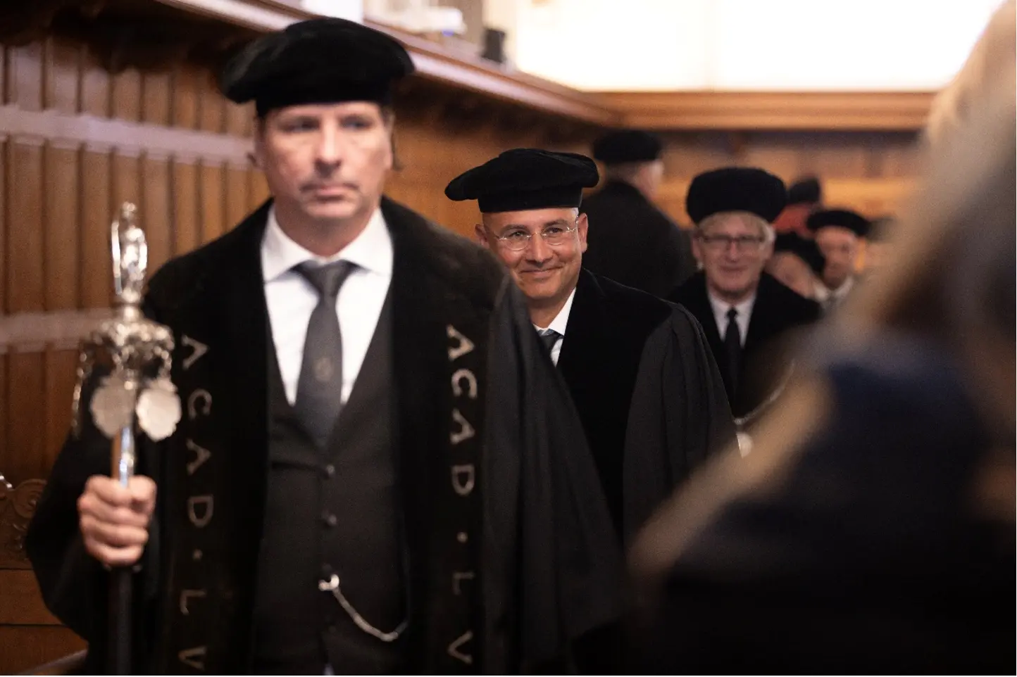 Accademici in abiti formali con berretti neri e mantelli, che sfilano in un ambiente interno elegante e tradizionale. Un uomo in primo piano tiene uno scettro, mentre altri sorridono sullo sfondo.