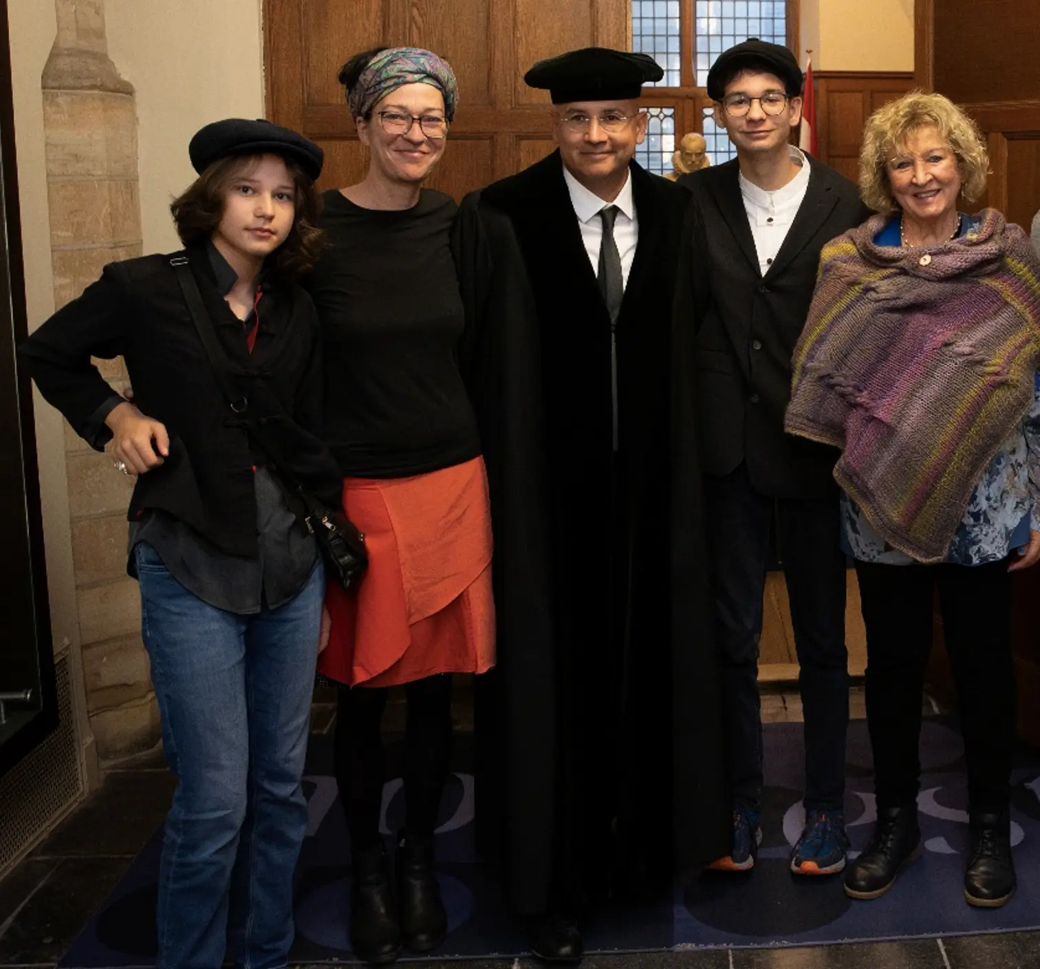 Un gruppo di cinque persone in abbigliamento formale, sorridenti e in piedi insieme in un ambiente interno elegante. Due persone indossano cappelli e quattro presentano abiti scuri, con una donna che indossa una gonna rossa e una sciarpa colorata.