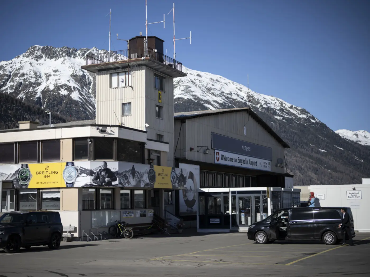 L'aeroporto di Engadin si trova in una giornata soleggiata, con montagne innevate sullo sfondo. La torre di controllo è visibile sulla sinistra, mentre sulla destra c'è l'ingresso principale con un cartello di benvenuto. Un veicolo parche