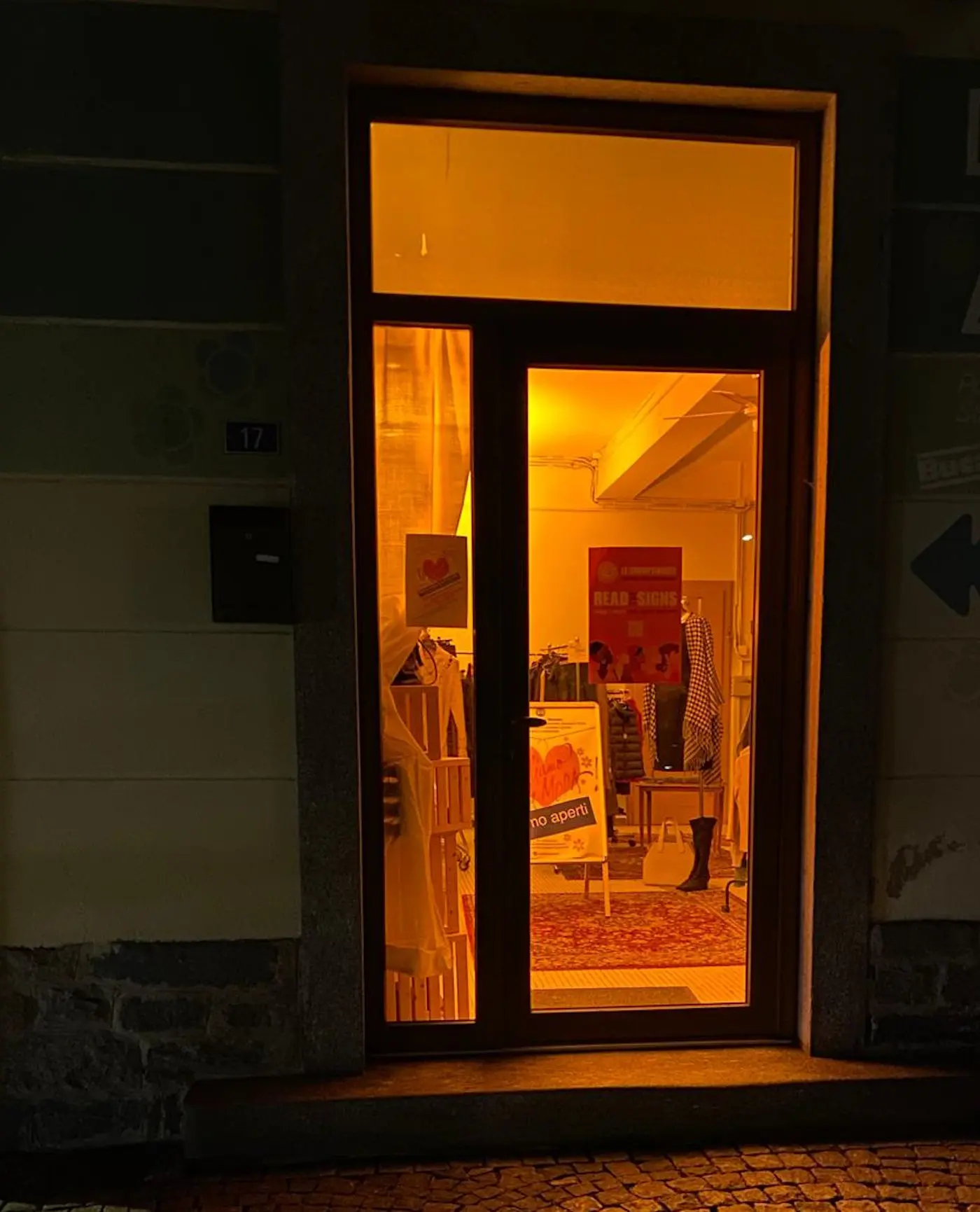 Una porta di ingresso illuminata da una luce arancione, che mostra l'interno di un negozio con abbigliamento esposto. All'interno si vedono diverse cose, tra cui un tappeto e cartelli, mentre l'esterno è buio.
