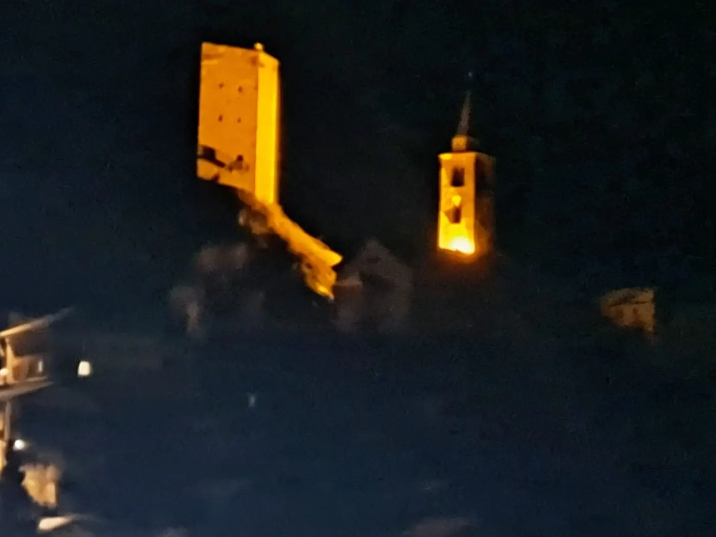 Ruine di un castello illuminate, con una torre campanaria visibile accanto. Scena notturna che evidenzia i contorni delle strutture storiche.
