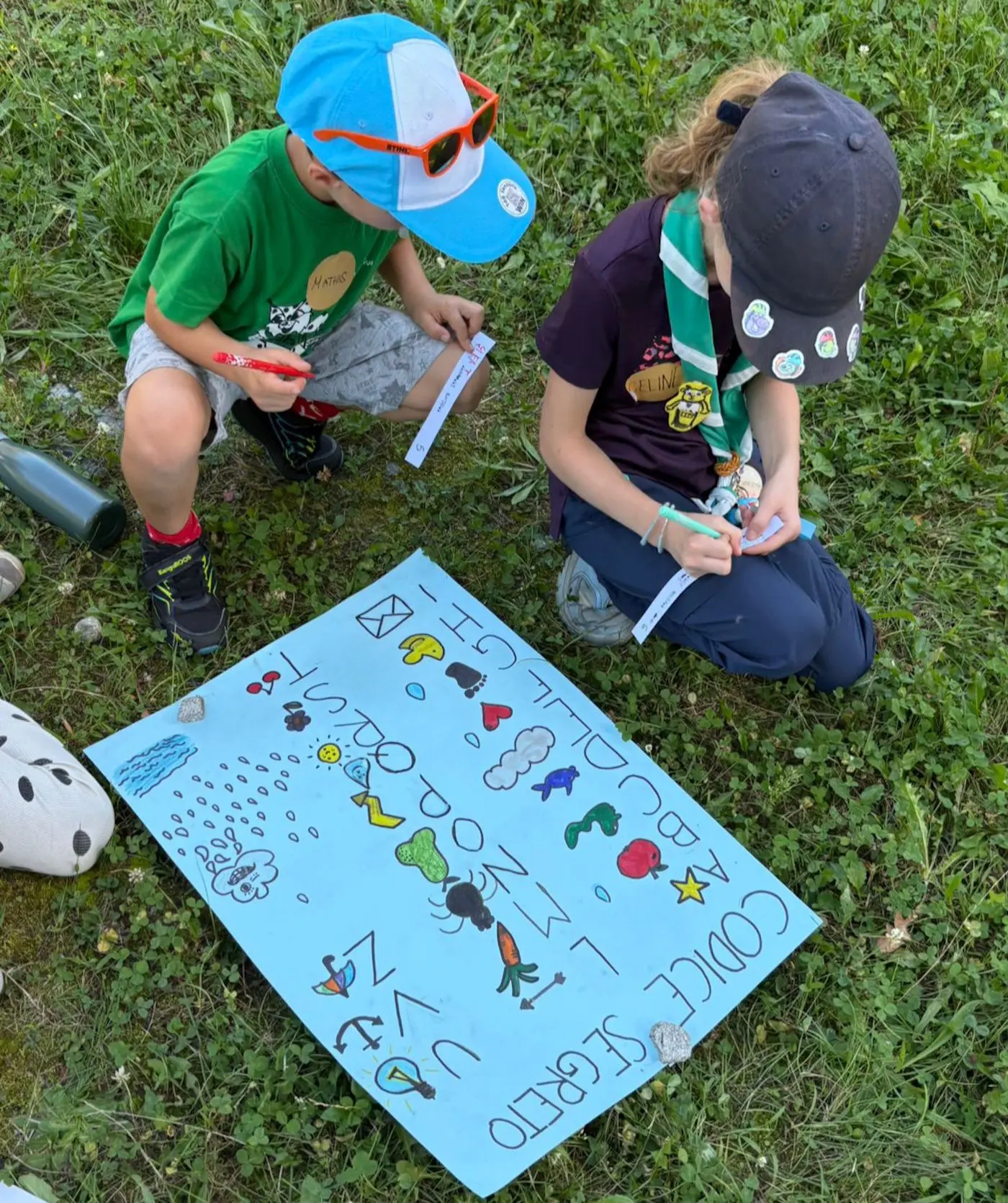 Due bambini seduti sull'erba, intenti a scrivere su un grande foglio di carta azzurra decorato con disegni di creature marine e lettere colorate. Uno indossa