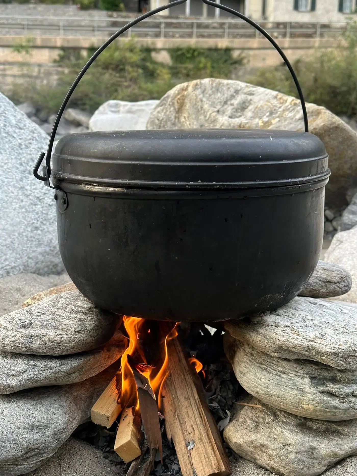 Un pentolone nero con manico è posto sopra un falò, sostenuto da rocce. Le fiamme ardono sotto il pentolone, circondato da ciottoli e natura
