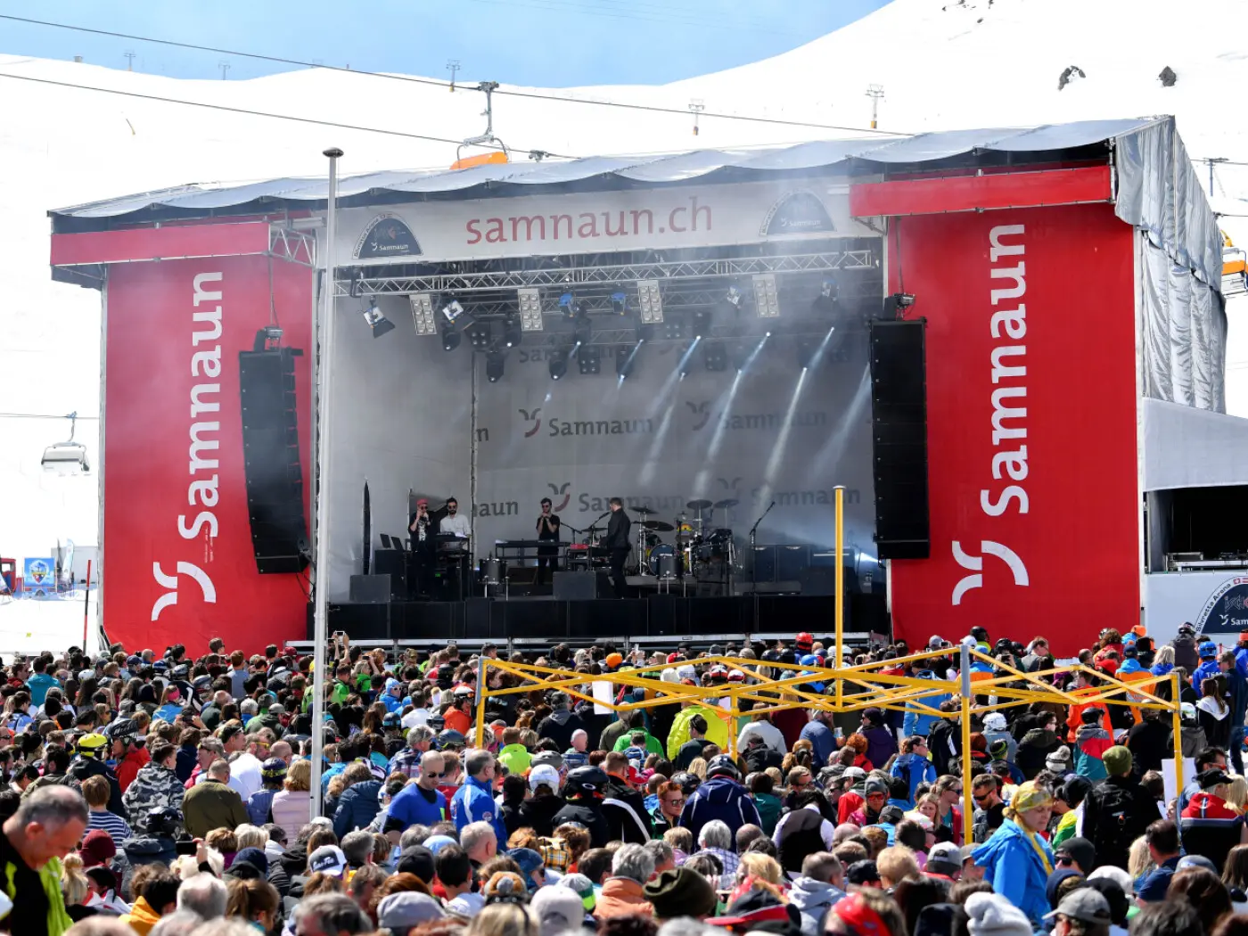 Un grande concerto all'aperto con migliaia di persone riunite davanti a un palco. Il palco è decorato con il logo "Sammaun" e circondato da luci