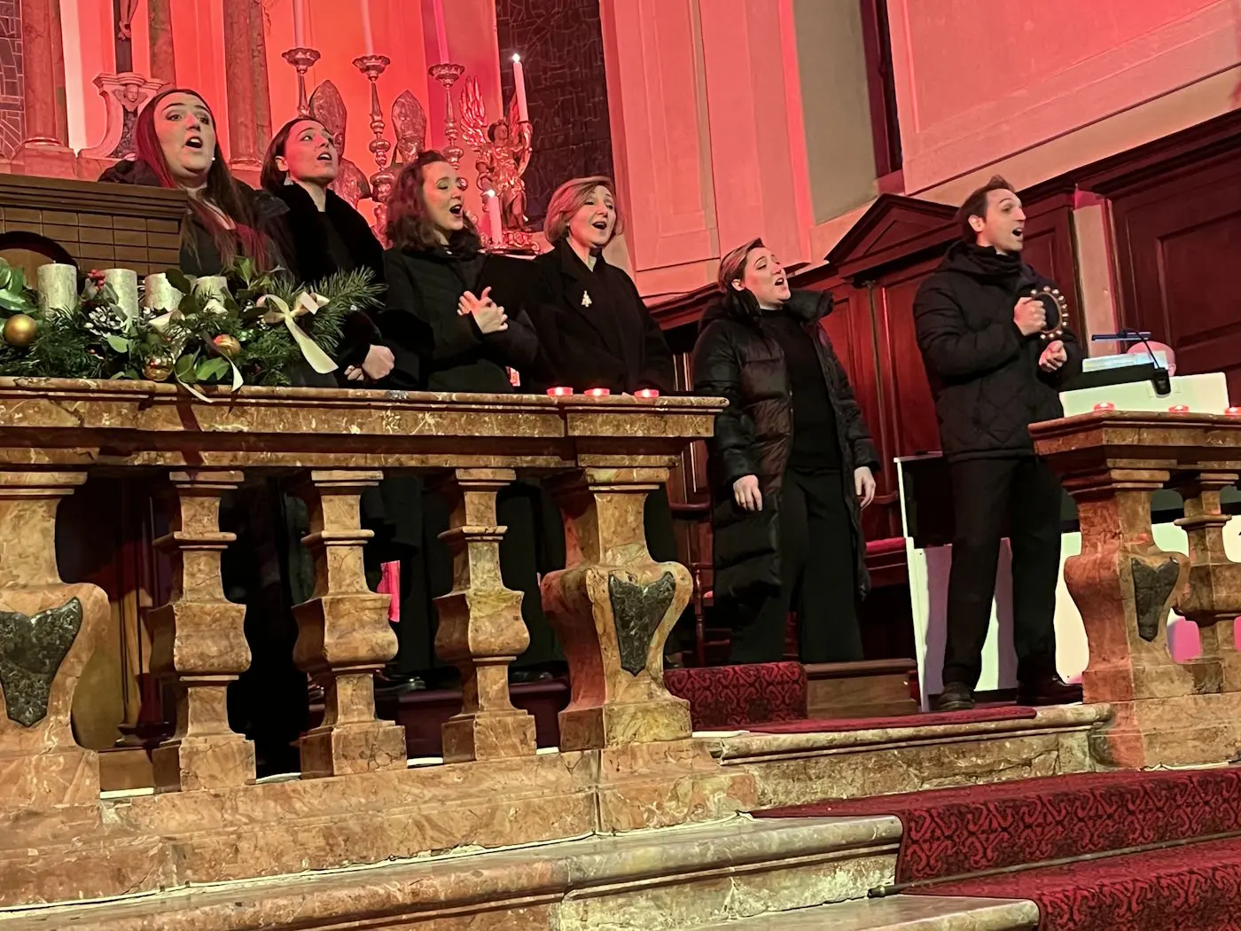 Un gruppo di cantanti in abbigliamento nero si esibisce su un palco decorato con candele e ornamenti natalizi. La scena è illuminata da luci rosse