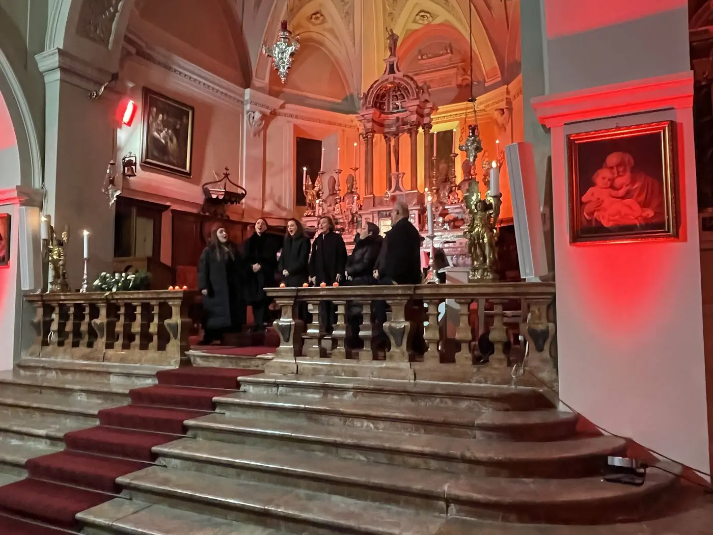 Un gruppo di persone in abiti scuri si trova su una balaustra all'interno di una chiesa illuminata con luci rosse e arancioni. Sullo sfondo si vede