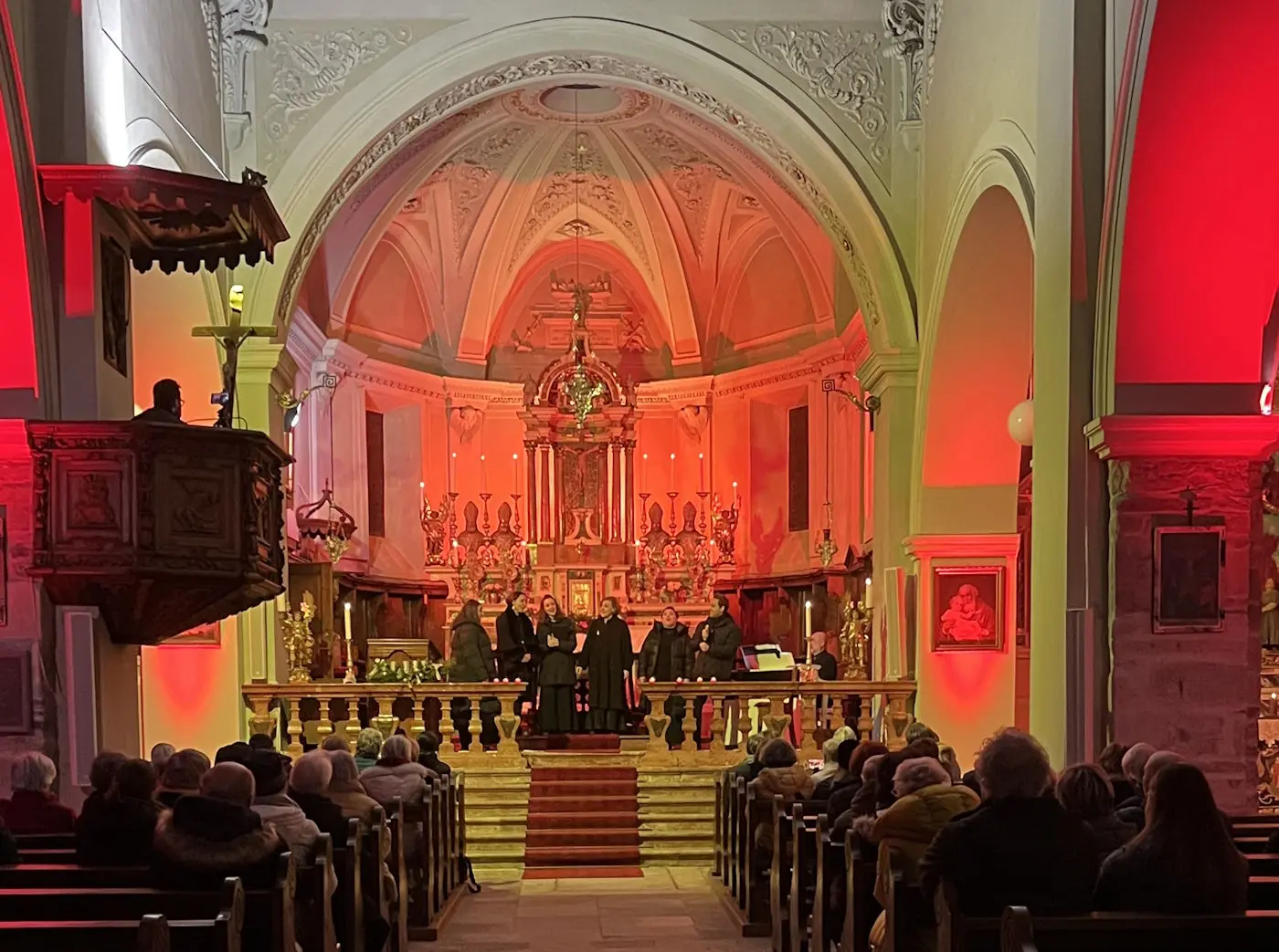 Interno di una chiesa con un'atmosfera illuminata da luci rosse e arancioni. Un gruppo di persone è sul palco, mentre il pubblico è seduto in diverse file