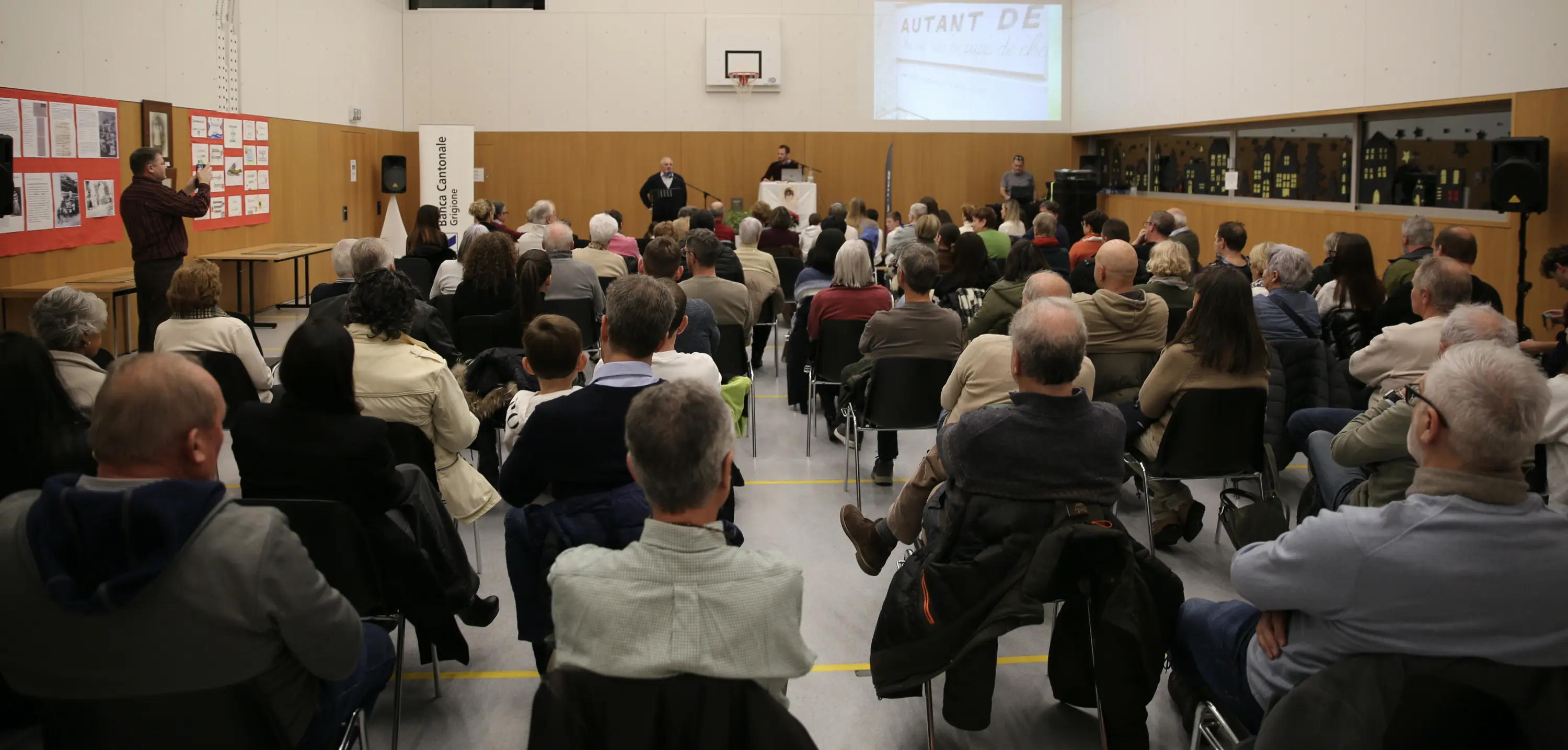 Una sala affollata con molte persone sedute che assistono a un evento. Sul palco, due relatori si trovano vicino a un microfono. Sullo sfondo, si vedono