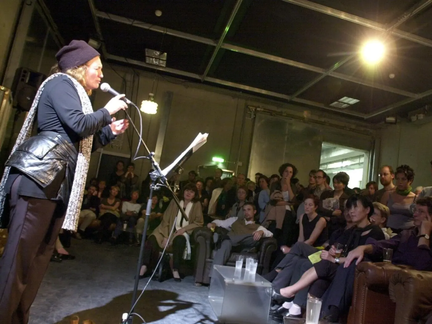 Una donna parla al microfono su un palco davanti a un pubblico attentamente radunato. La sala è buia, mentre una luce illumina la relatrice. Il pubblico, seduto su