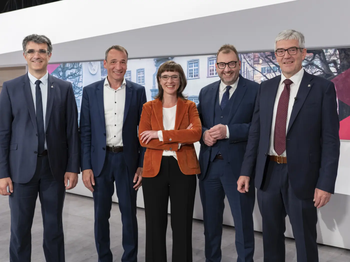 Cinque persone in abiti formali sorridono mentre posano insieme su un palco. Sullo sfondo, uno schermo con immagini storiche.