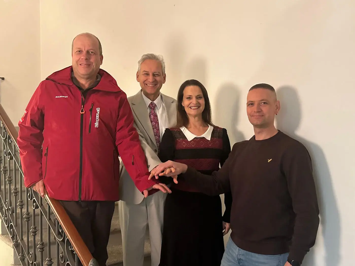 Quattro persone sorridenti posano insieme su una scala. Un uomo in giacca rossa è accanto a un uomo in abito chiaro, mentre una donna in abito nero e una