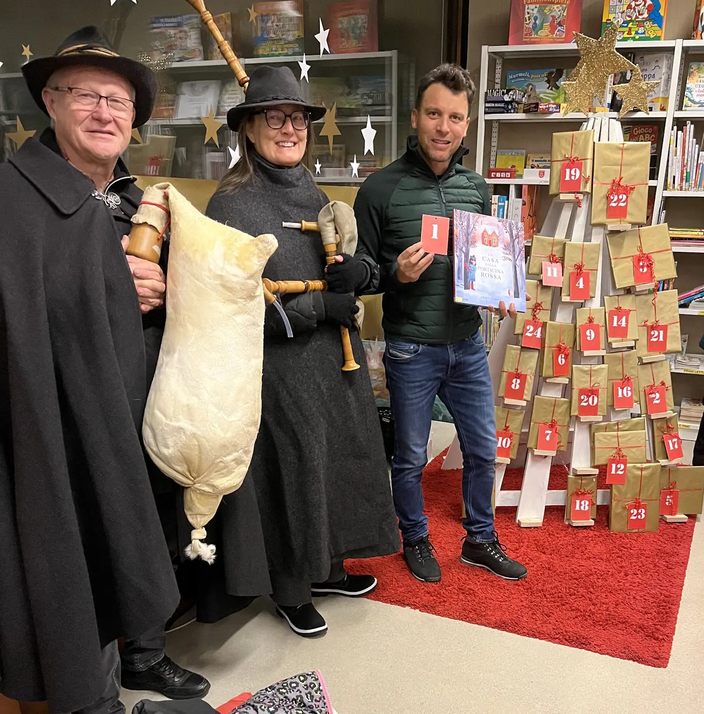 Tre persone sorridenti, vestite in abiti festivi, si trovano accanto a un albero decorato con sacchetti di carta numerati. Uno tiene un libro, mentre gli