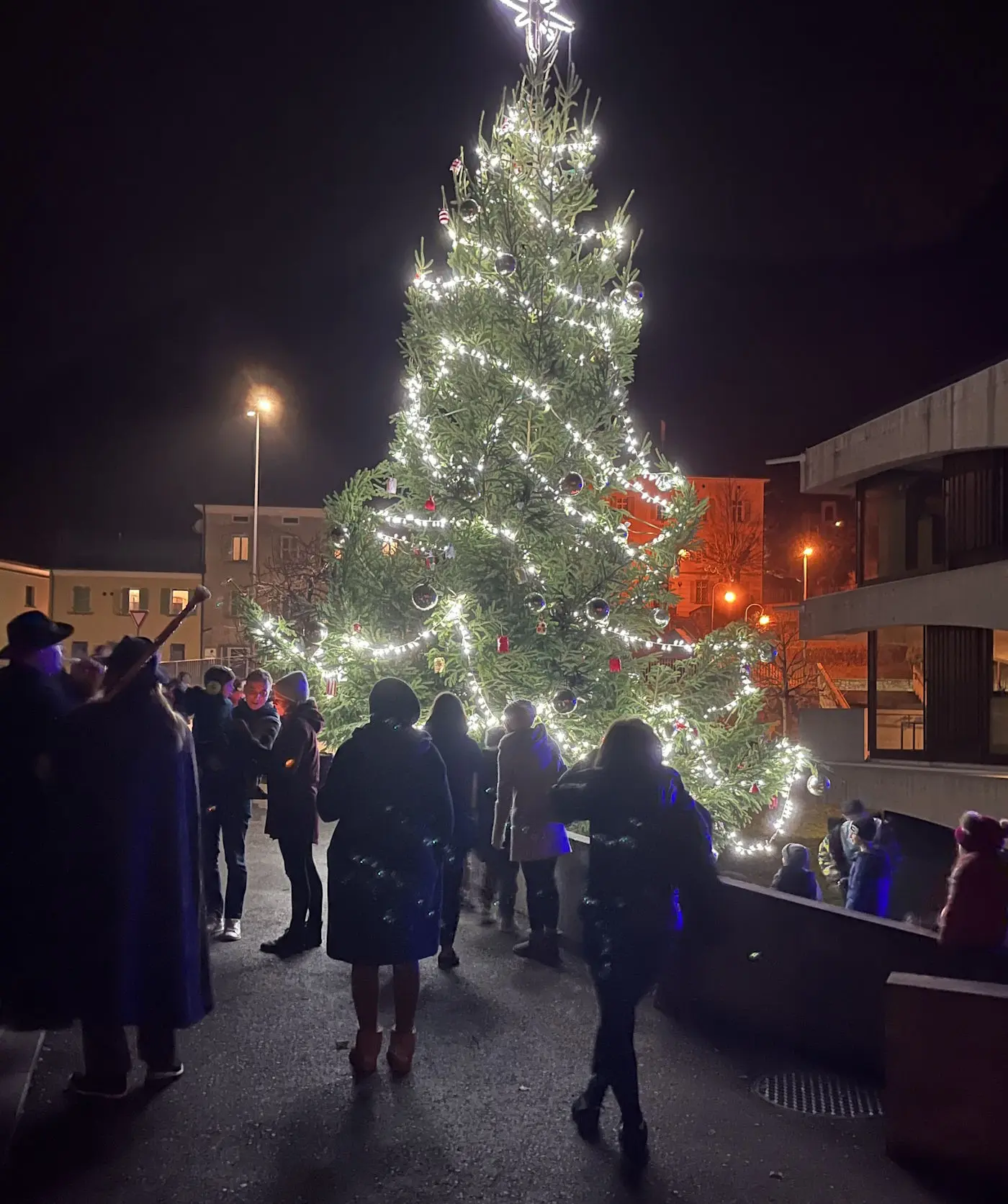 Albero di Natale illuminato con decorazioni, circondato da persone durante un evento festivo notturno. Luci brillanti e atmosfera gioiosa.