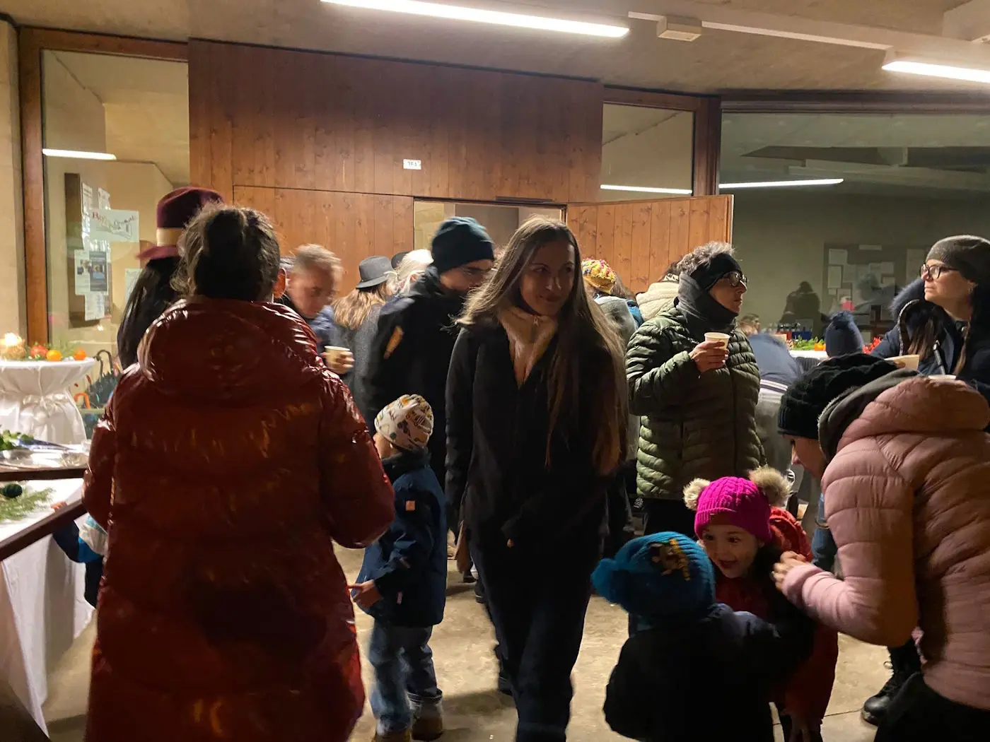 Una folla di persone in un ambiente festivo, con adulti e bambini che interagiscono. Alcuni indossano giacche e cappelli invernali, mentre altri sorse